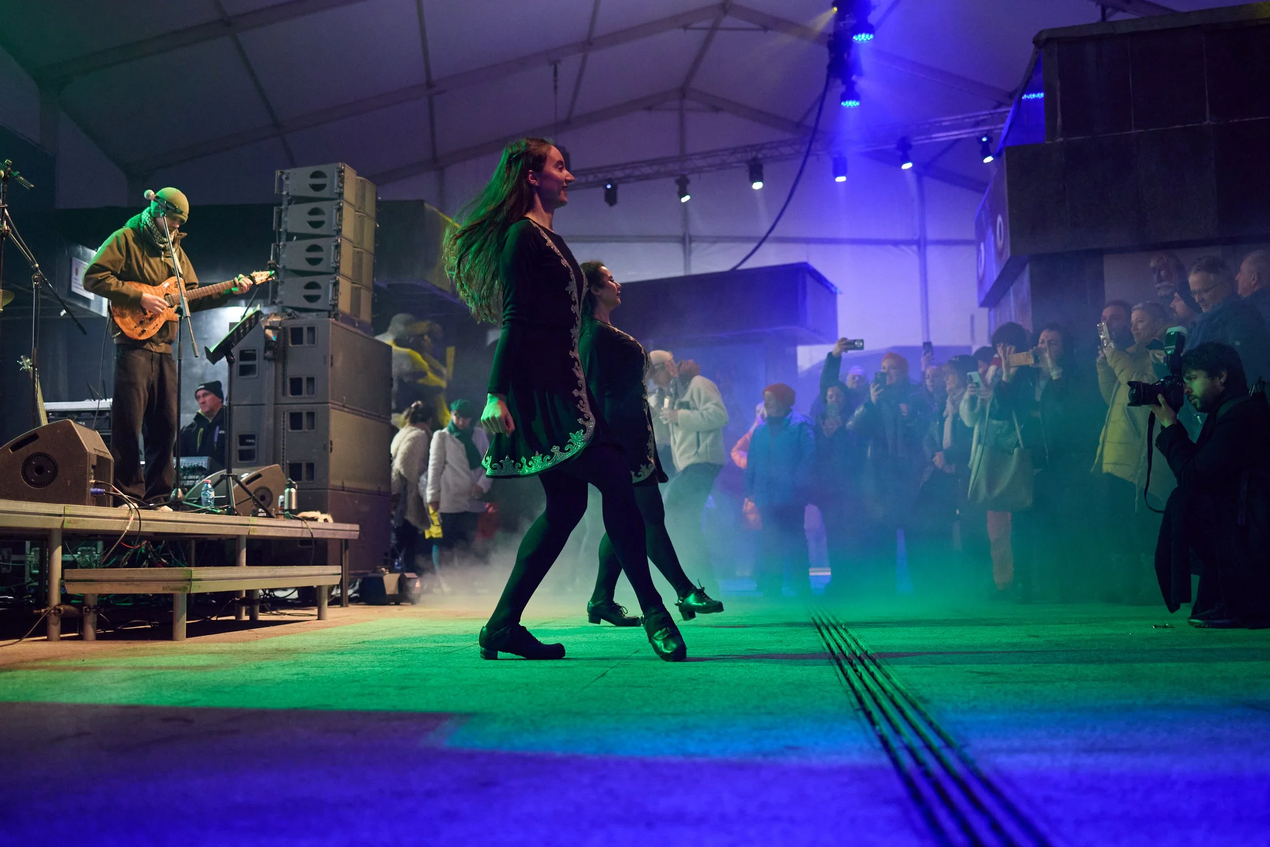 Two women in black dance shoes and black tights perform on a stage illuminated with colorful lights, while a crowd watches, some taking photos, in a large indoor venue.