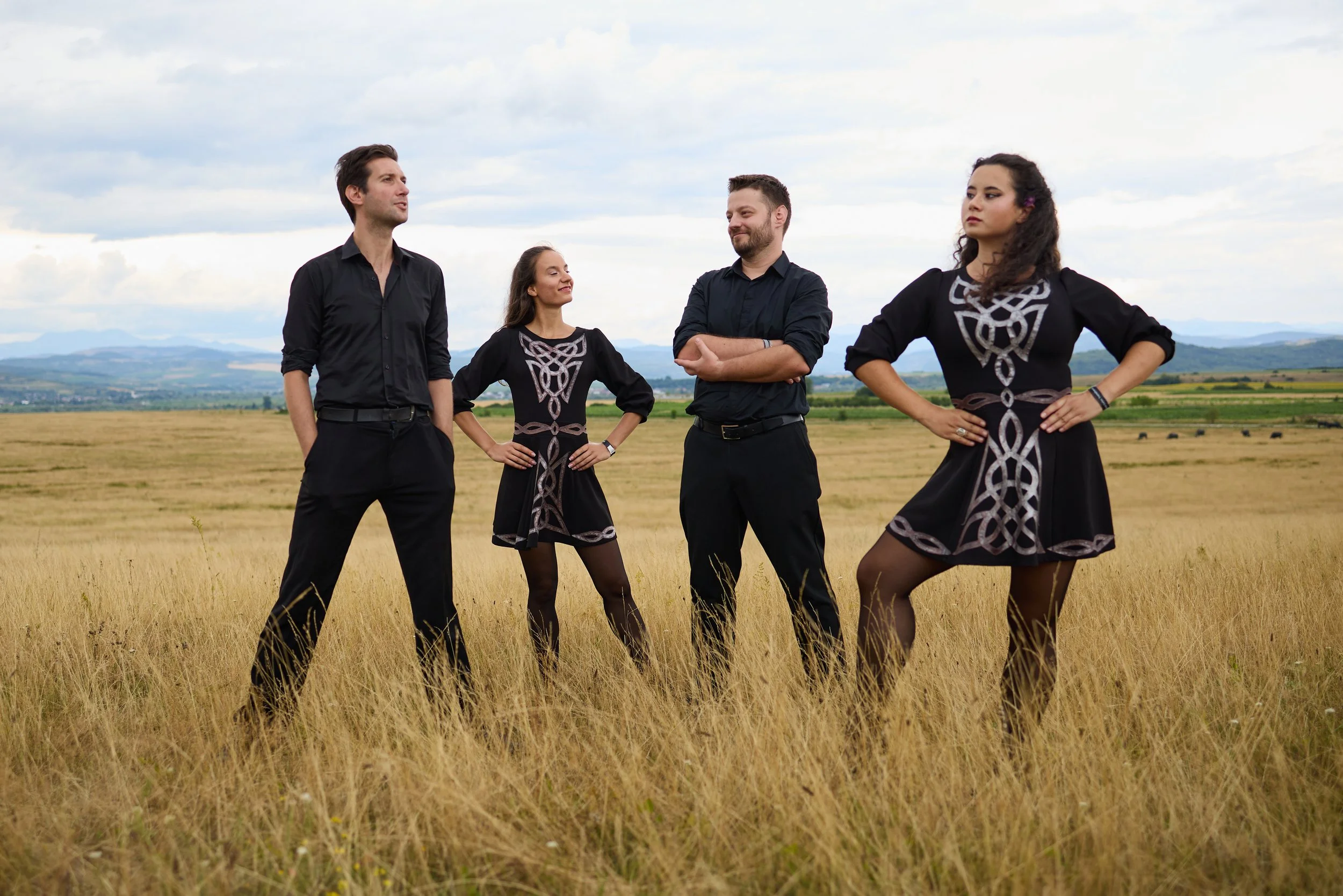 Four people standing in a wheat field with mountains and cloudy sky in the background. Two women and two men, dressed in black with Celtic designs, posing confidently.