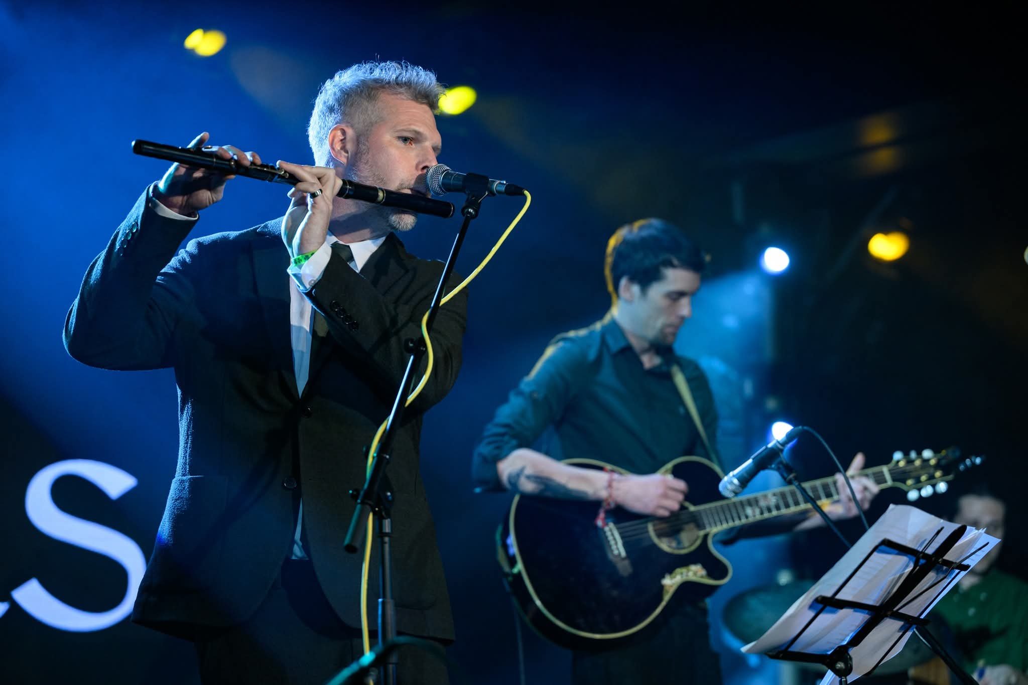 Two musicians performing on stage, one playing the flute and the other playing the guitar, with stage lights in the background.