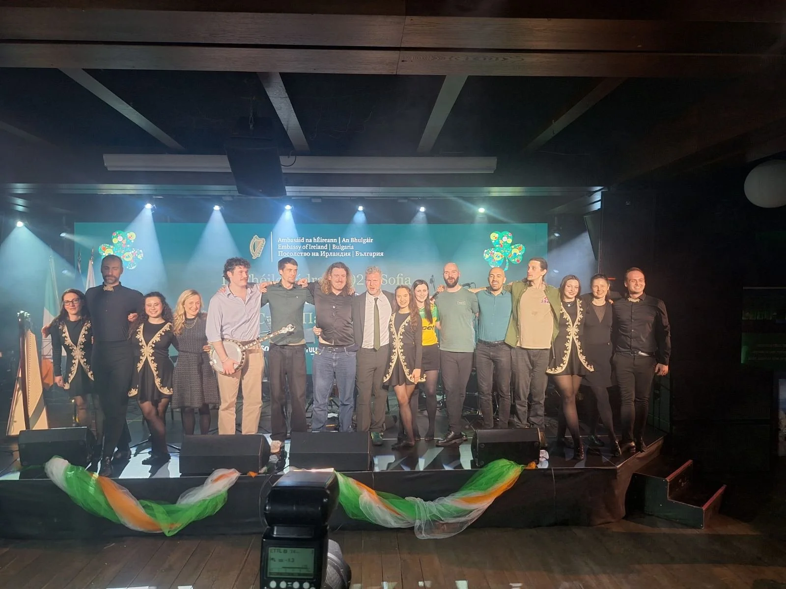 A group of people on stage at an event, standing together and smiling, with a backdrop displaying text in multiple languages and logos, lit by stage lights, with some holding musical instruments.