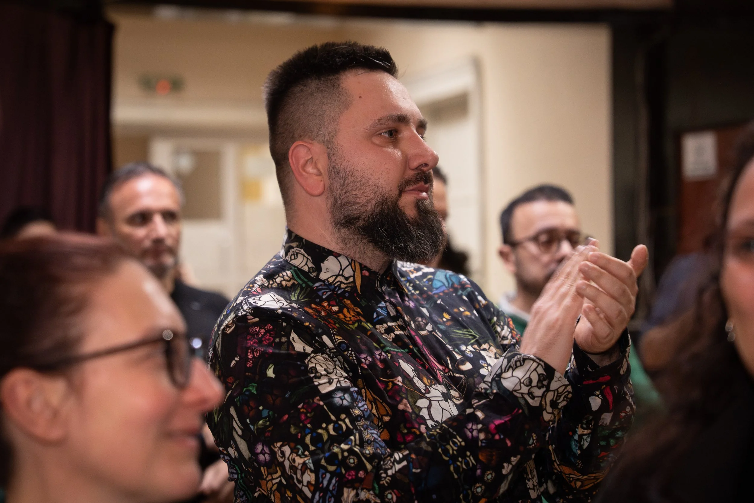 A man with a beard and short dark hair wearing a colorful, patterned shirt claps during an indoor event, surrounded by people.