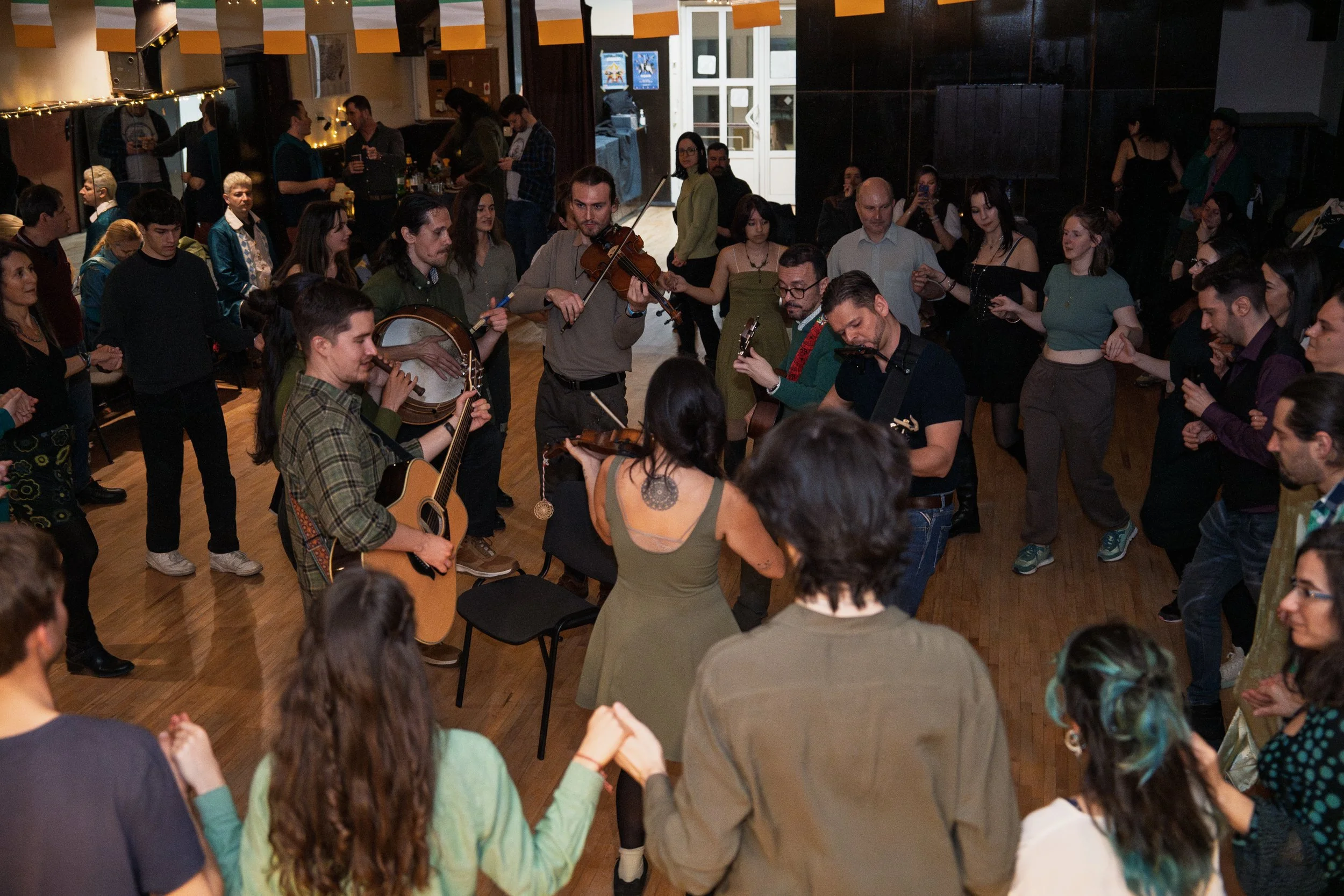 A group of people dancing in a circle at a party or celebration, with musicians playing instruments in the center, in a warmly lit indoor setting.