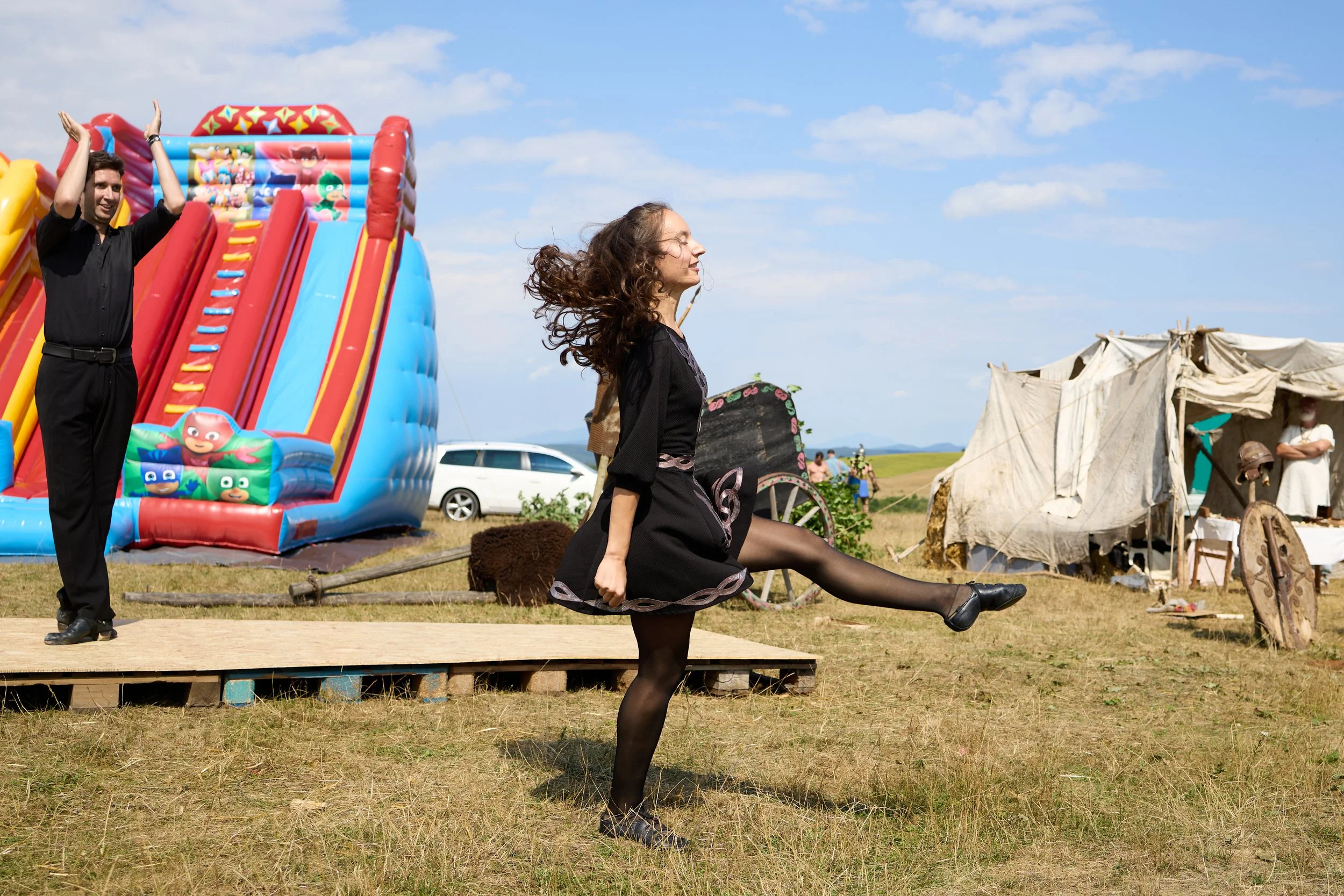 A woman dressed in a black dress and tights performing a high kick in front of a colorful inflatable slide at an outdoor event. A man in black clothes stands behind her with his hands raised, smiling. There are tents and people in the background, sug