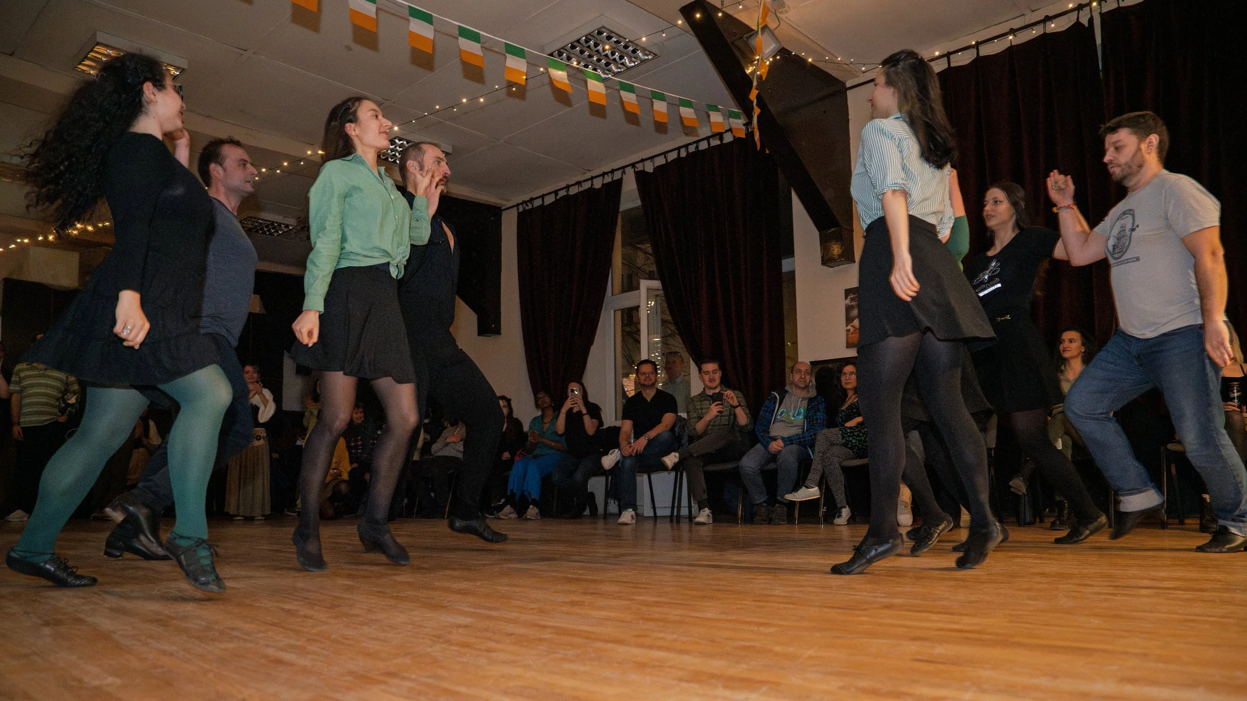A group of people dancing together on a wooden floor in front of an audience in a dimly lit room decorated with Irish flags and string lights.