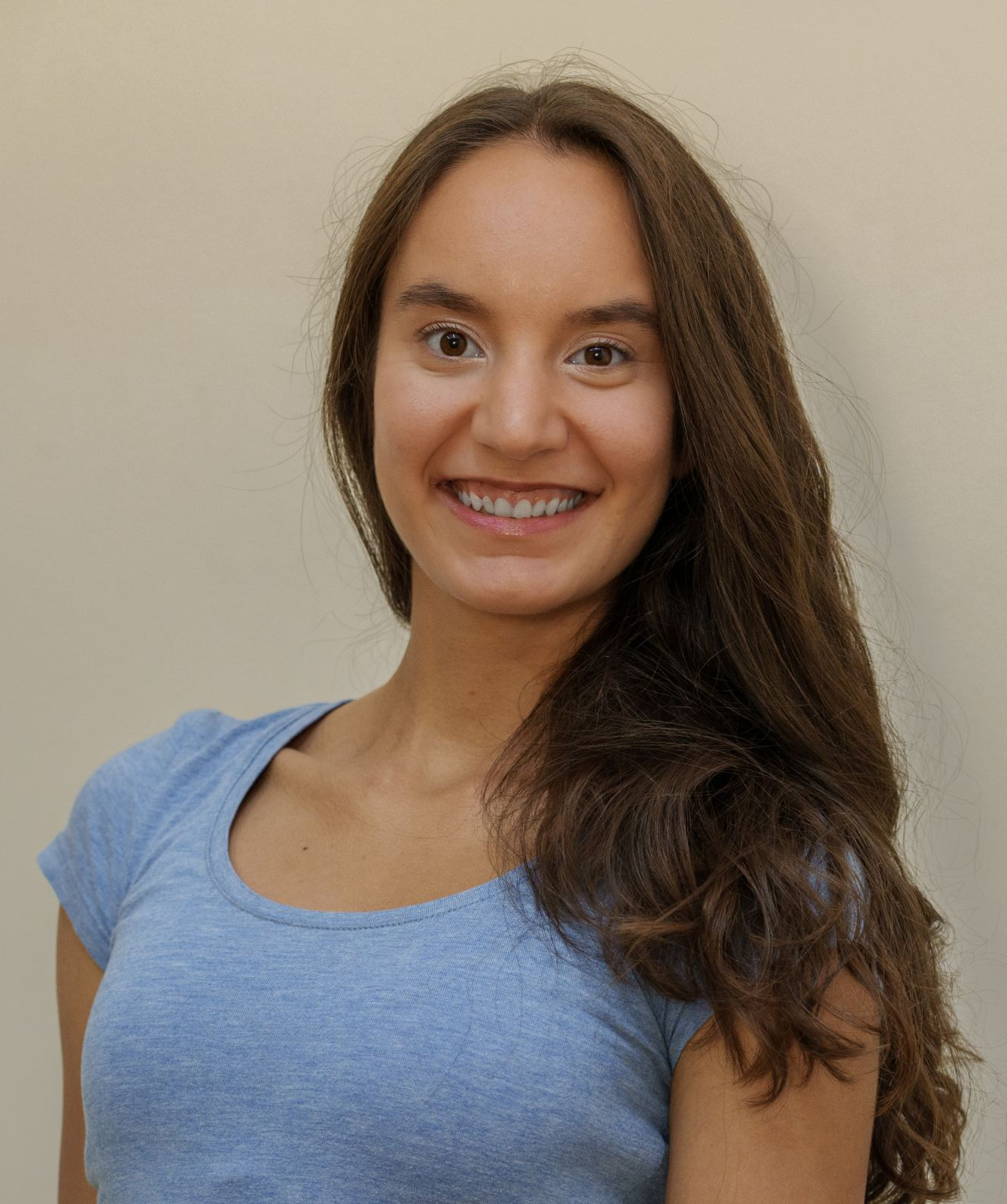 Headshot of a young woman with long, wavy brown hair, smiling and wearing a light blue T-shirt against a plain background.