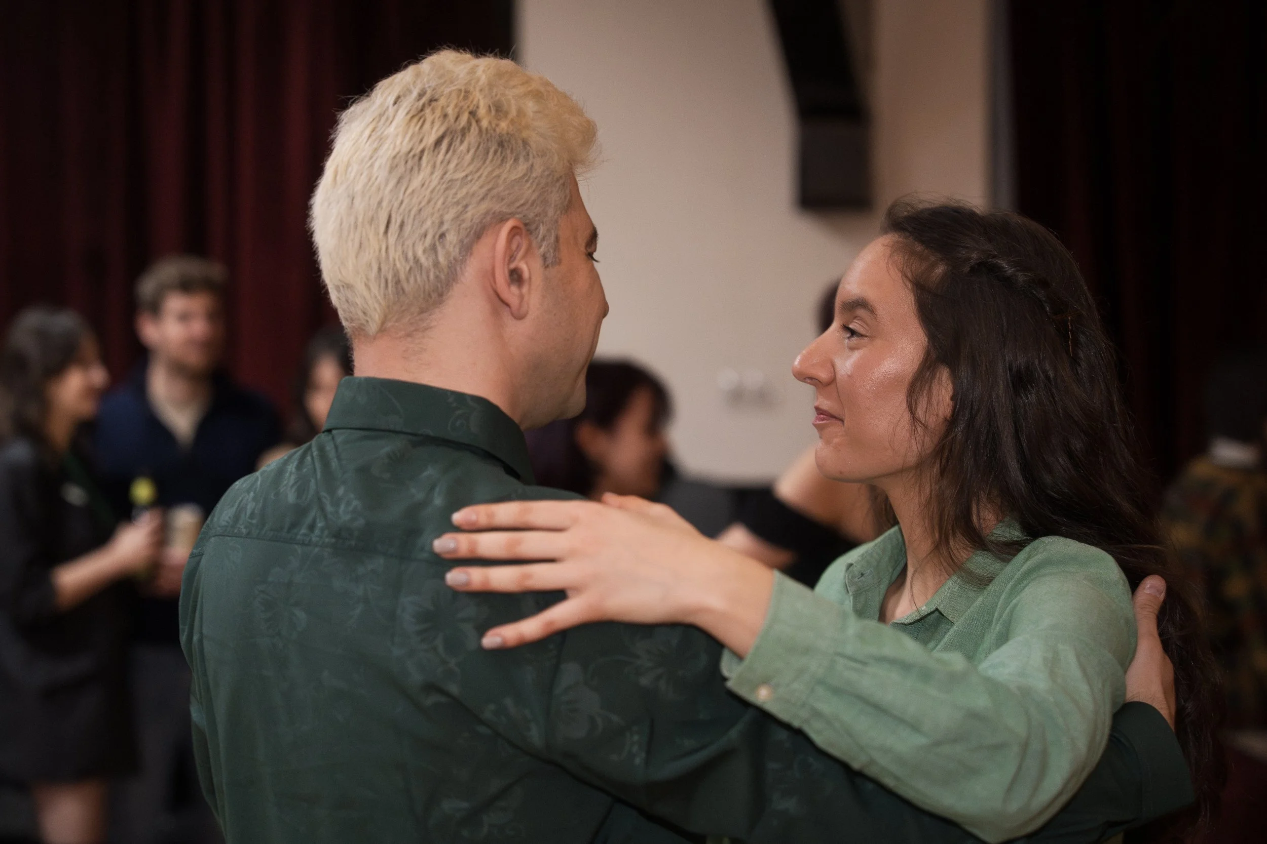 A man with platinum blonde hair and a woman with dark wavy hair dancing closely together indoors, with other people in the background, possibly at a social event or dance class.
