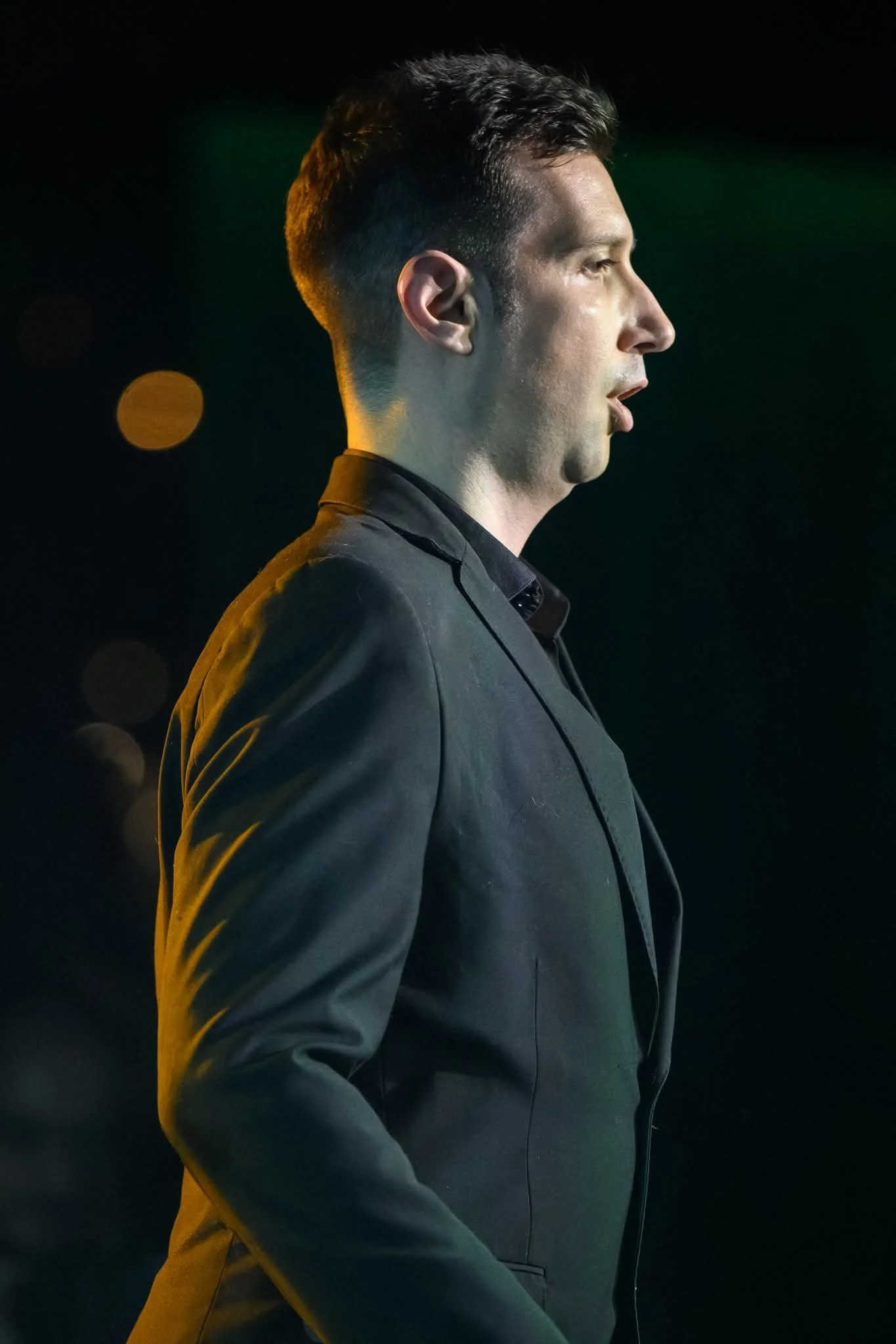 Profile view of a man in a dark suit against a dark background with bokeh lights, illuminated by side lighting.