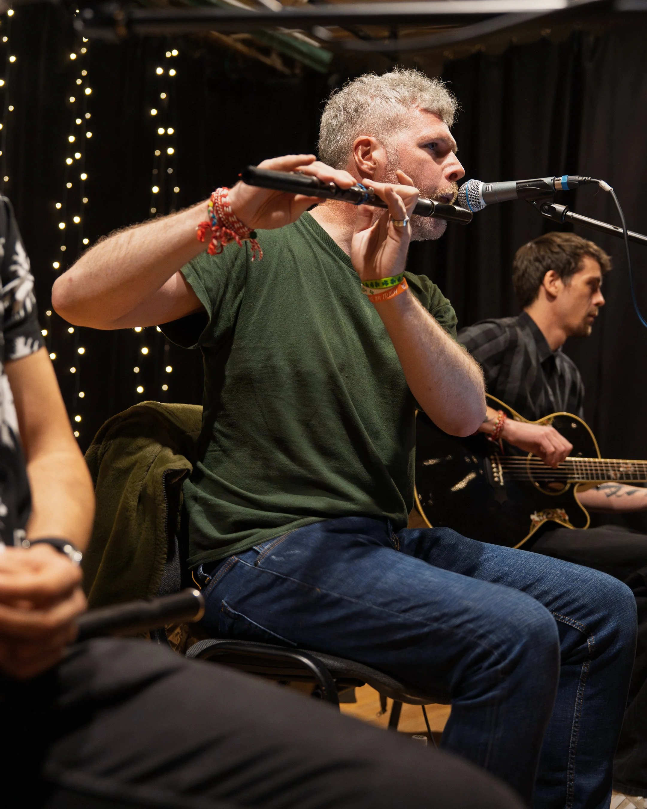 A man with gray hair and a beard playing a flute during a musical performance, with another man with dark hair and a guitar sitting beside him. The background has black curtains with string lights.