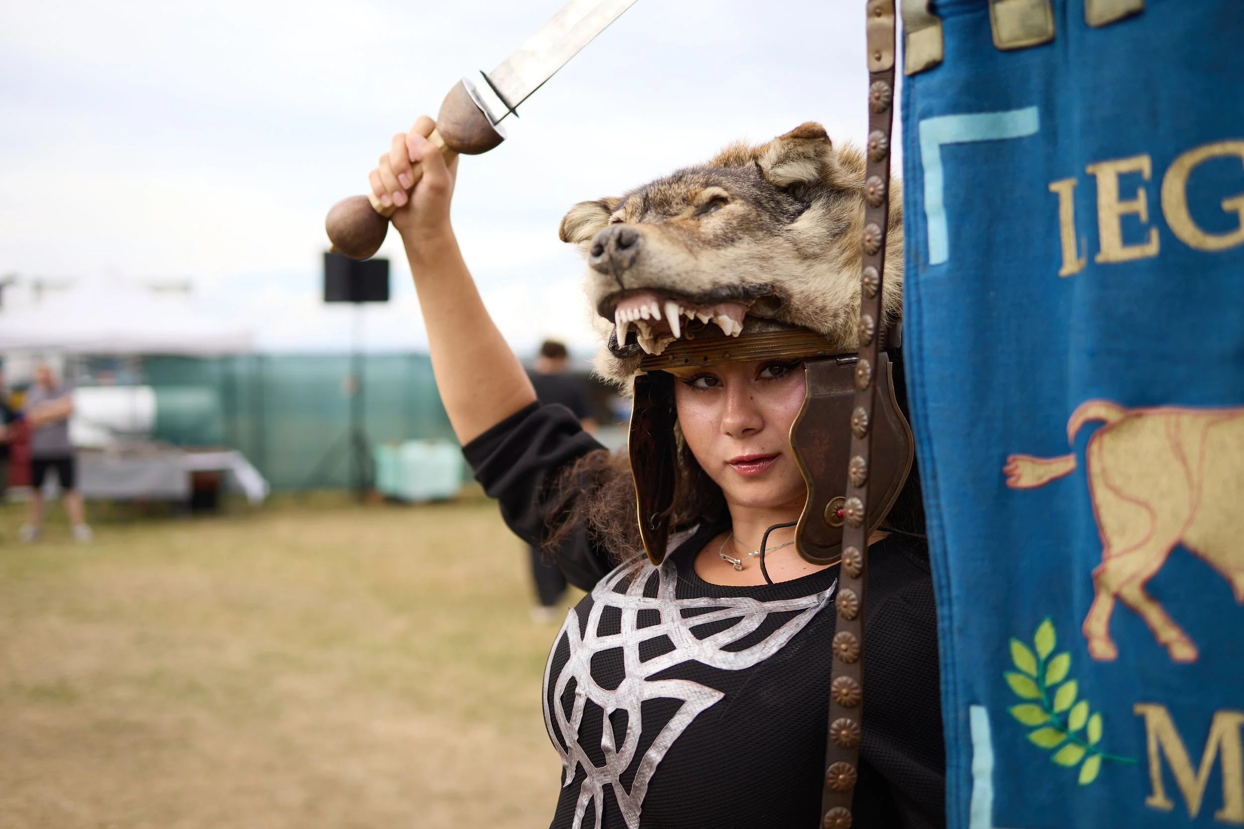 A woman dressed as a medieval knight wearing a lion's head helmet and holding a sword at a festival or reenactment event.