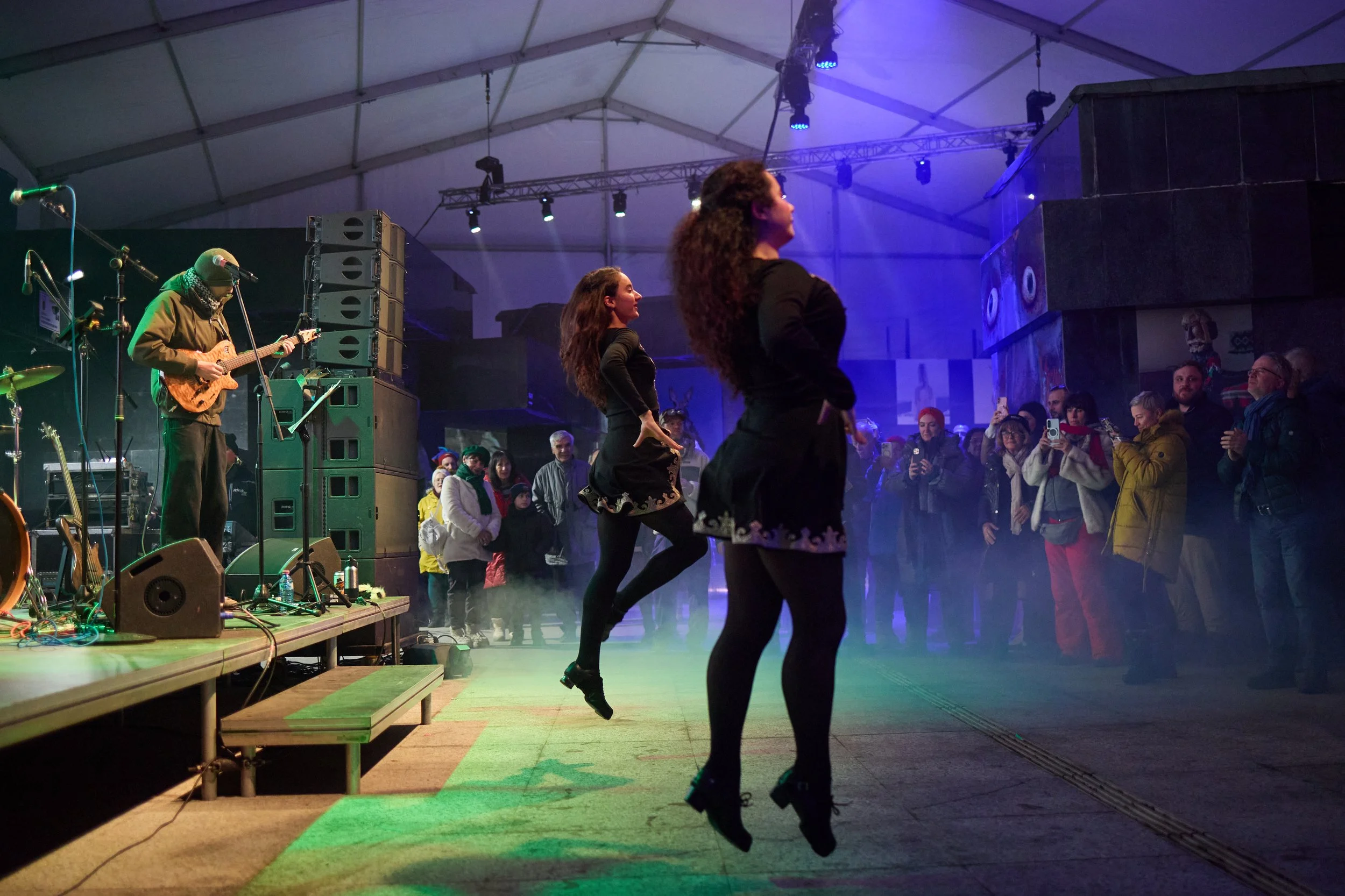 Two women dancing in black dresses on a green-lit stage while a band plays with one man playing guitar. Audience members watch the performance inside a large tent with stage lighting.