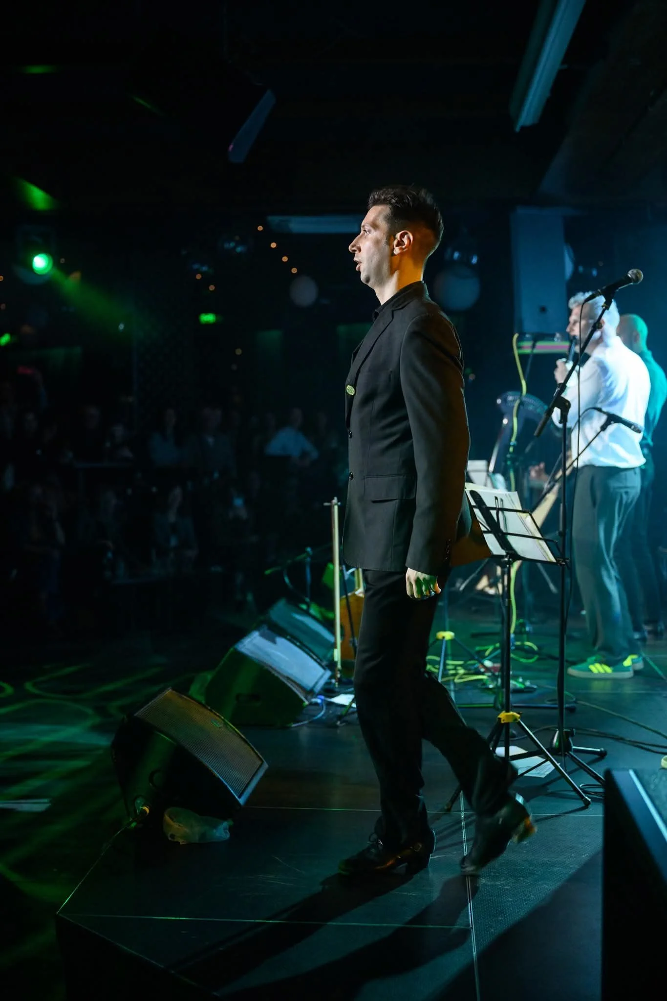 A man with dark hair and a black suit standing on stage with a microphone, musical equipment, and other performers in the background, under stage lighting.