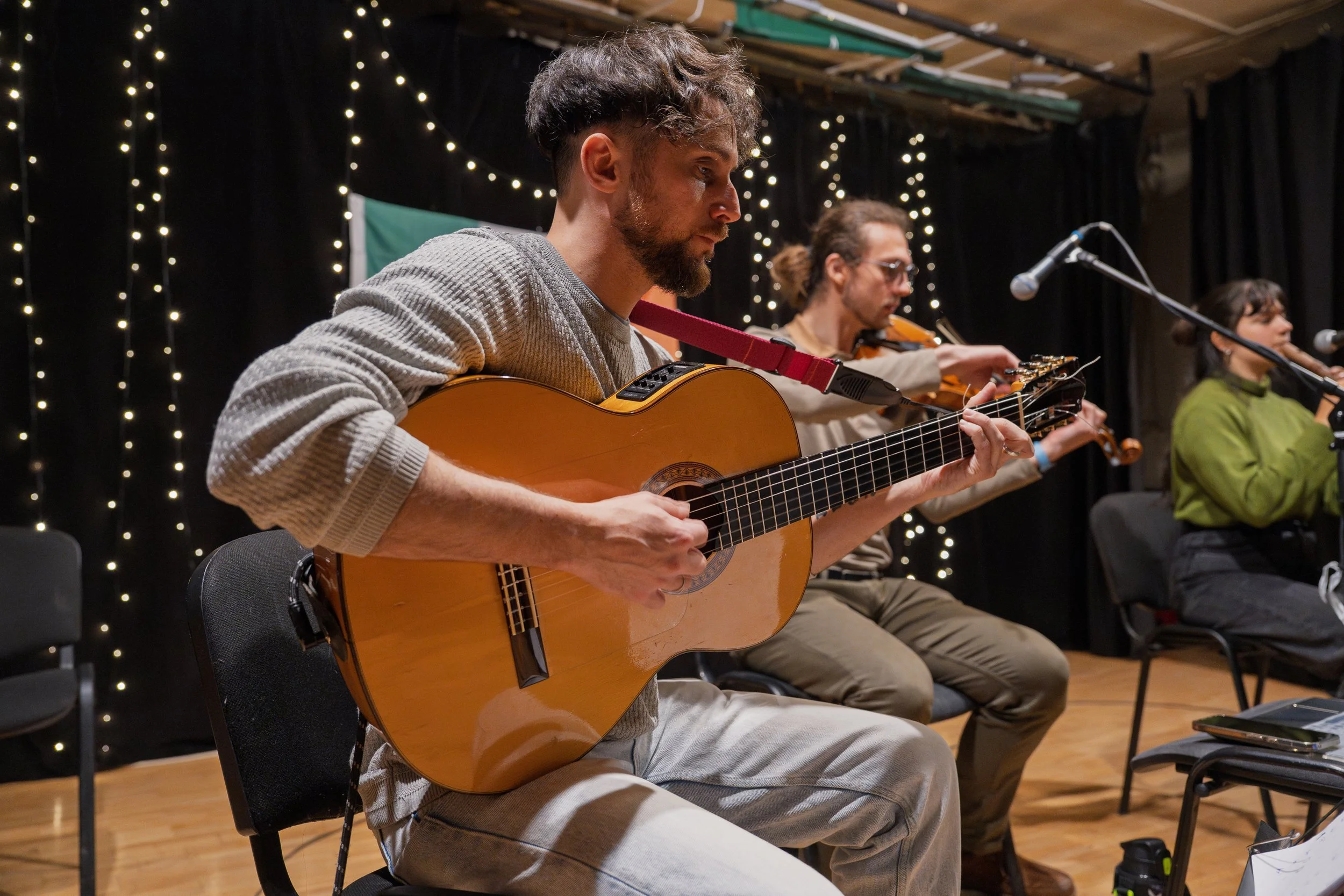 A man playing an acoustic guitar during a musical performance in a dimly lit indoor venue with string lights and a black curtain background, accompanied by other musicians.