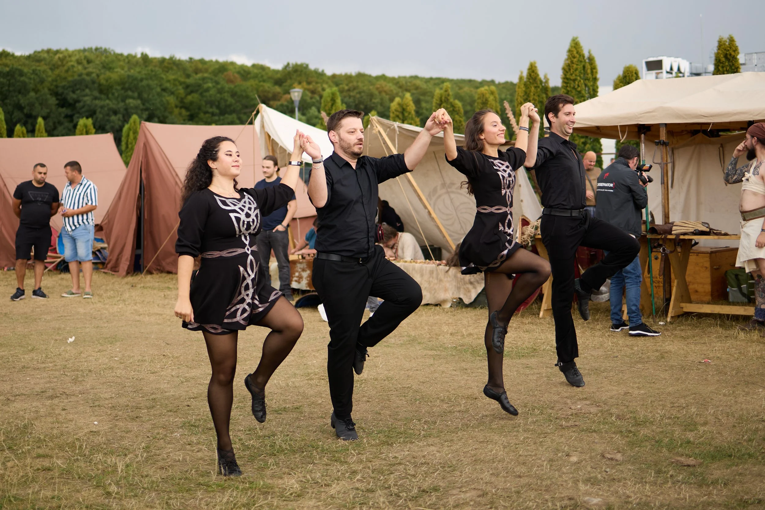 Group of four people dancing outdoors in a grassy area with tents and trees in the background.