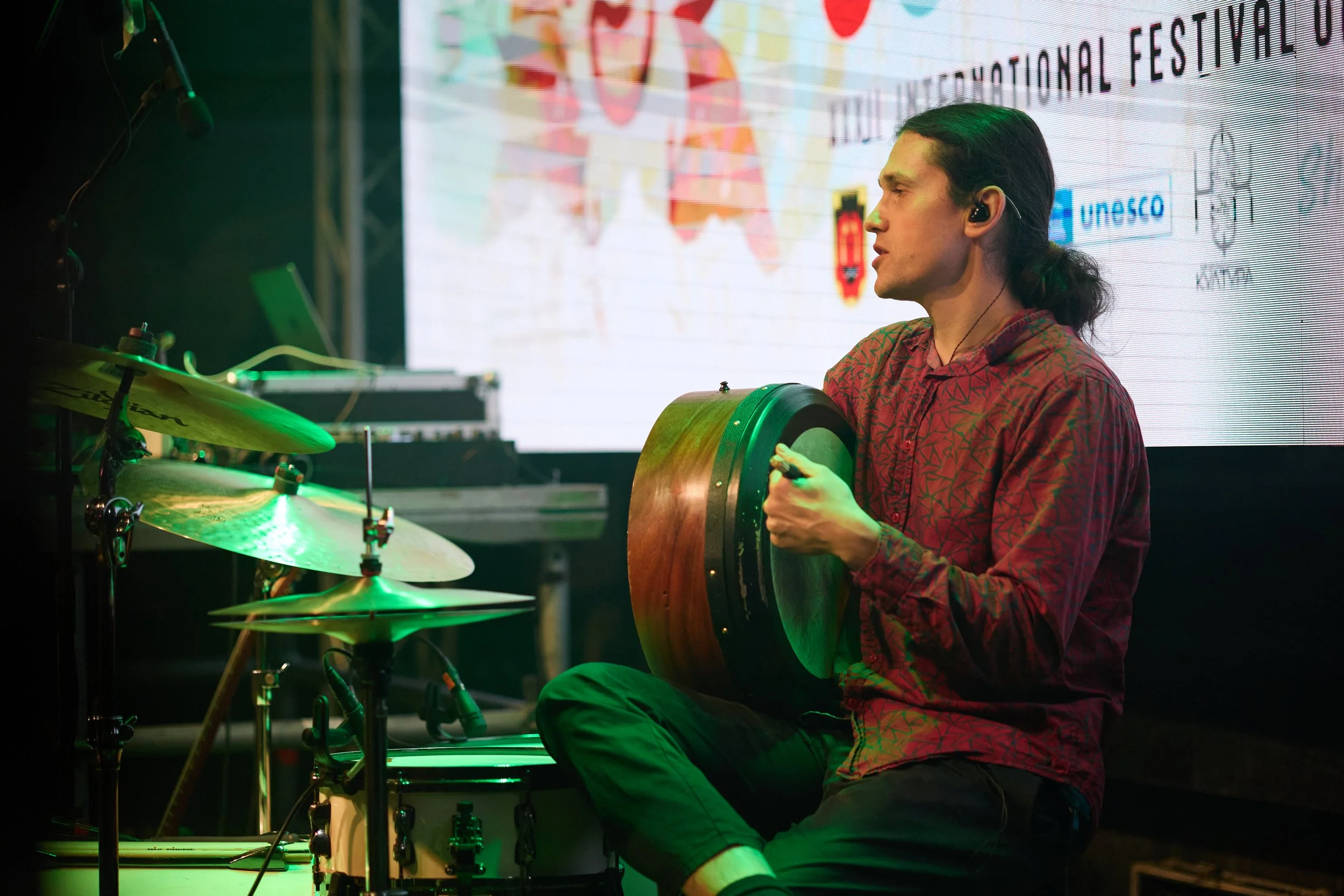 A young man with long dark hair playing drums and holding a percussion instrument on stage during a performance or event.