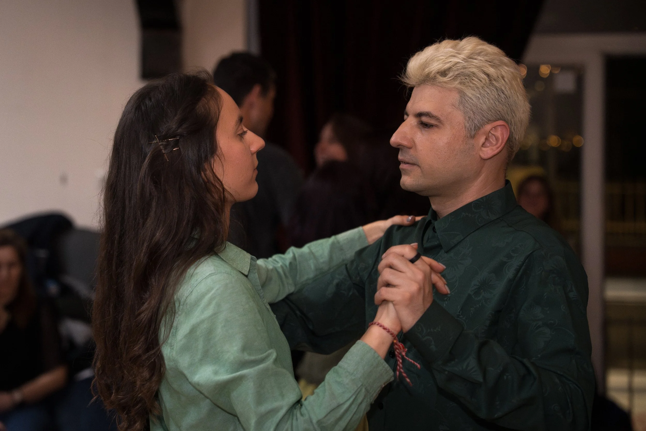 A man and woman are dancing closely, holding hands, and gazing into each other's eyes in an indoor setting with dim lighting and other people in the background.