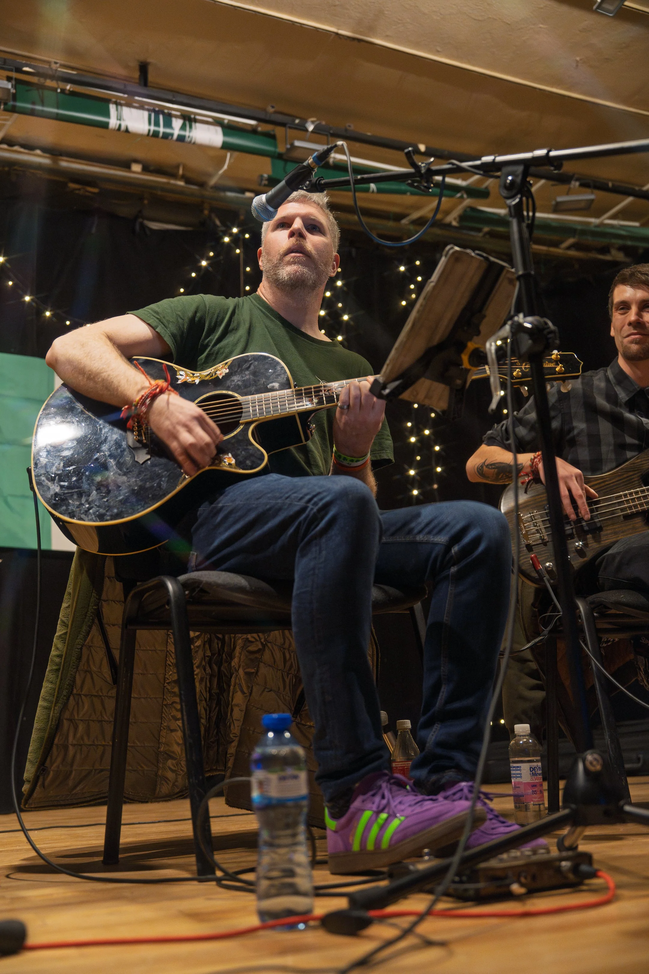 Musicians performing with guitars on stage, with microphones, music stands, and lights in the background.