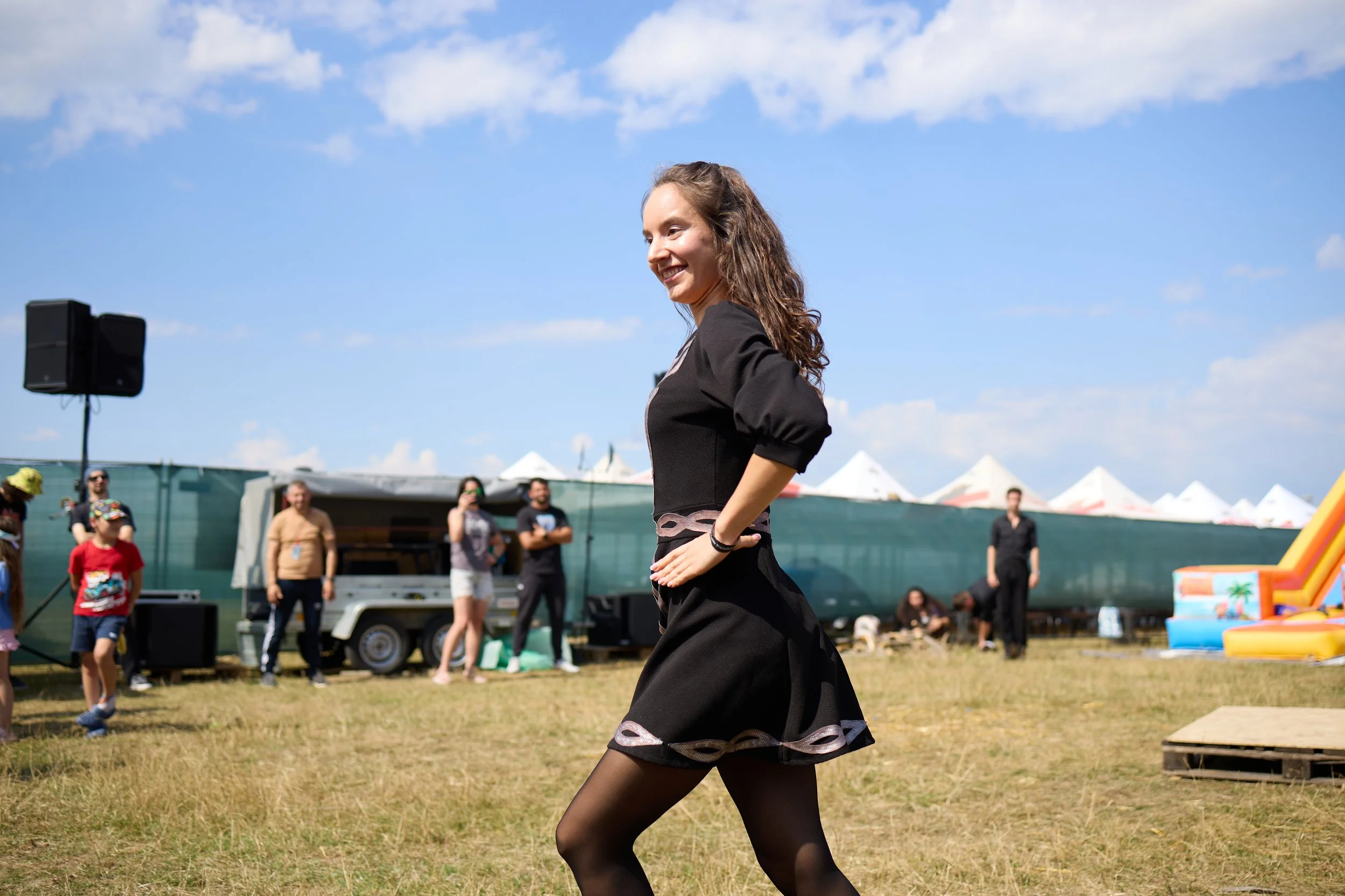 A young woman in ballet attire dancing outdoors at a festival with a crowd watching under a partly cloudy blue sky.