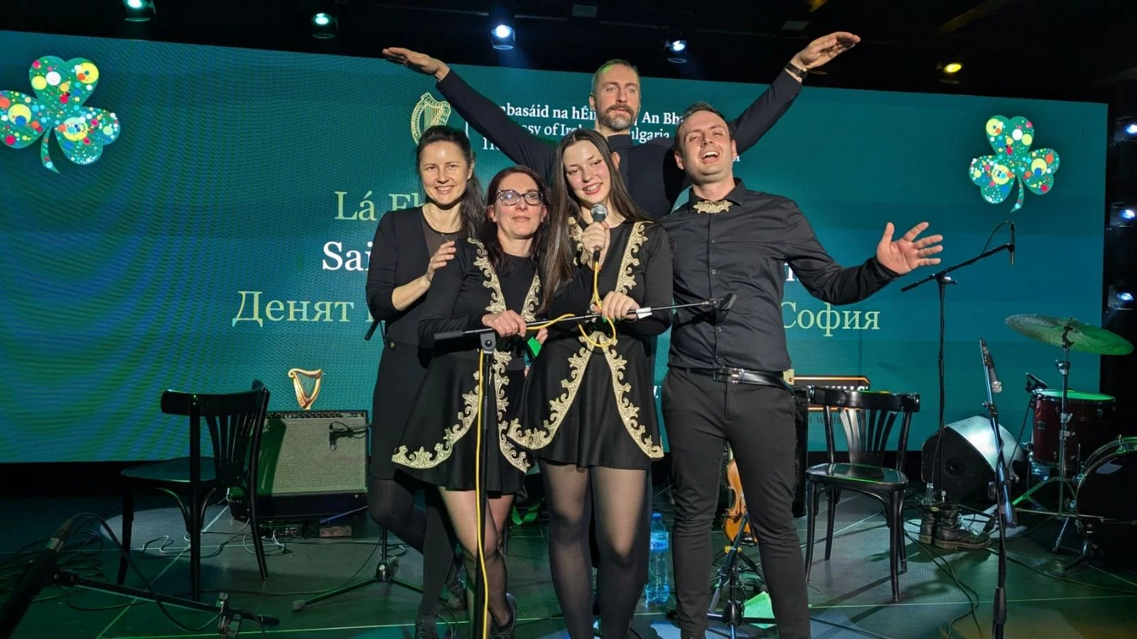 Group of five musicians on stage during a performance, with a large green screen behind them displaying text in various languages, musical instruments including a harp, guitar, and drums, and stage equipment.