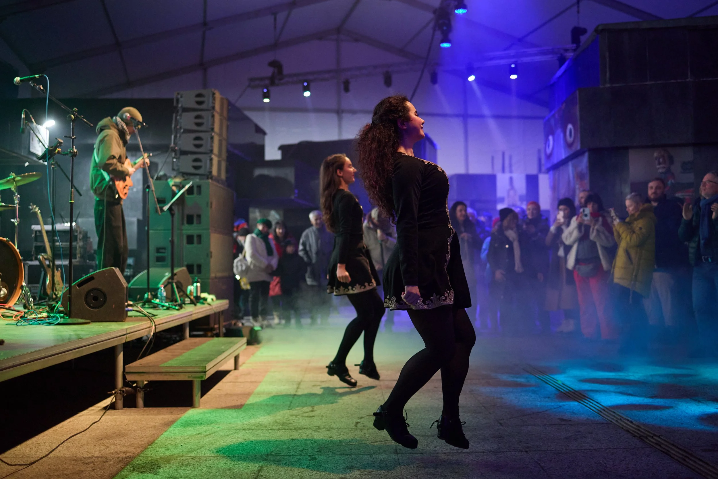 Women dancing on a stage at a live music event with musicians playing instruments in the background and an audience watching.