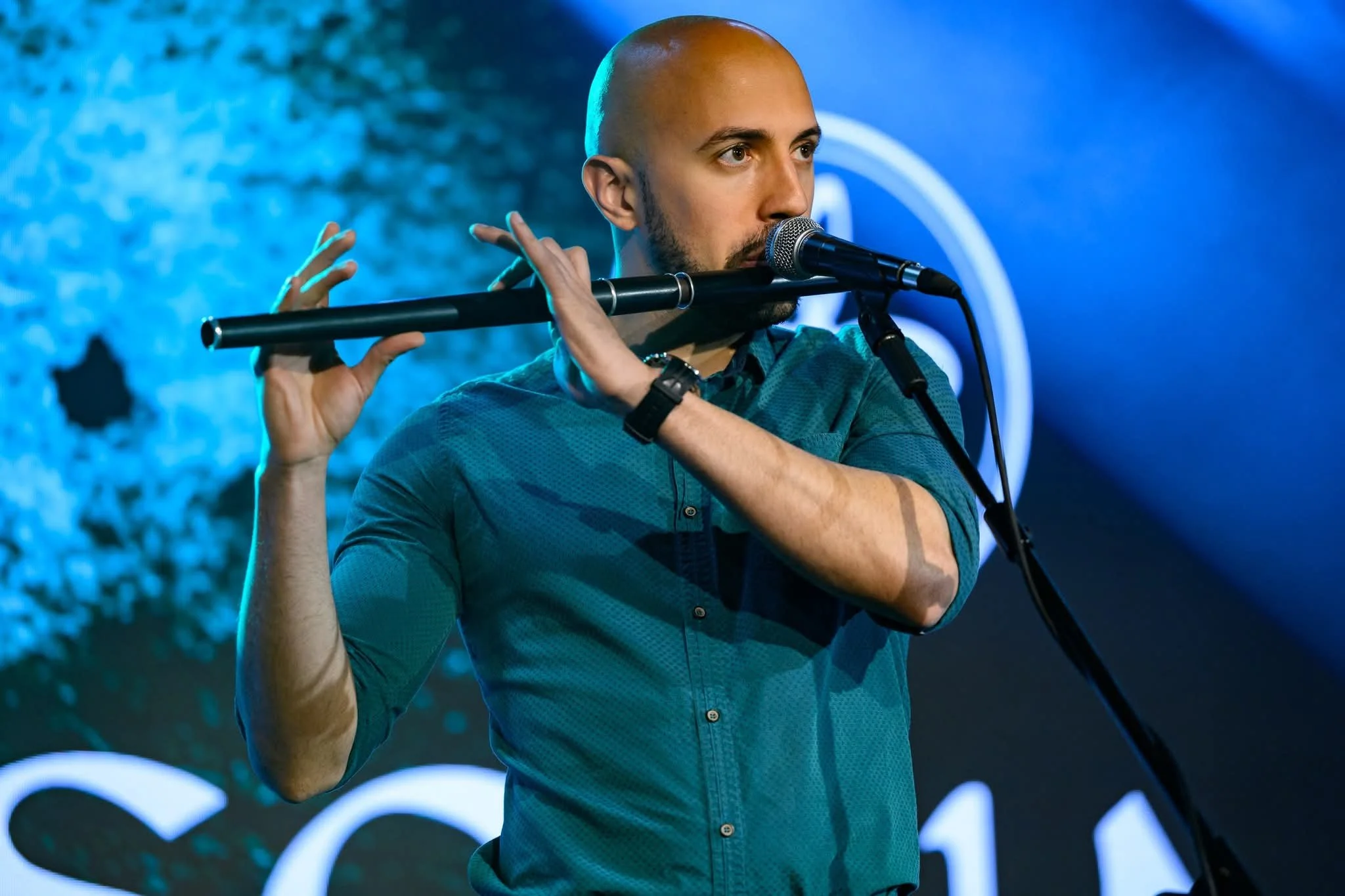 A man with a shaved head and beard is playing a flute in front of a blue background with decorative lighting.