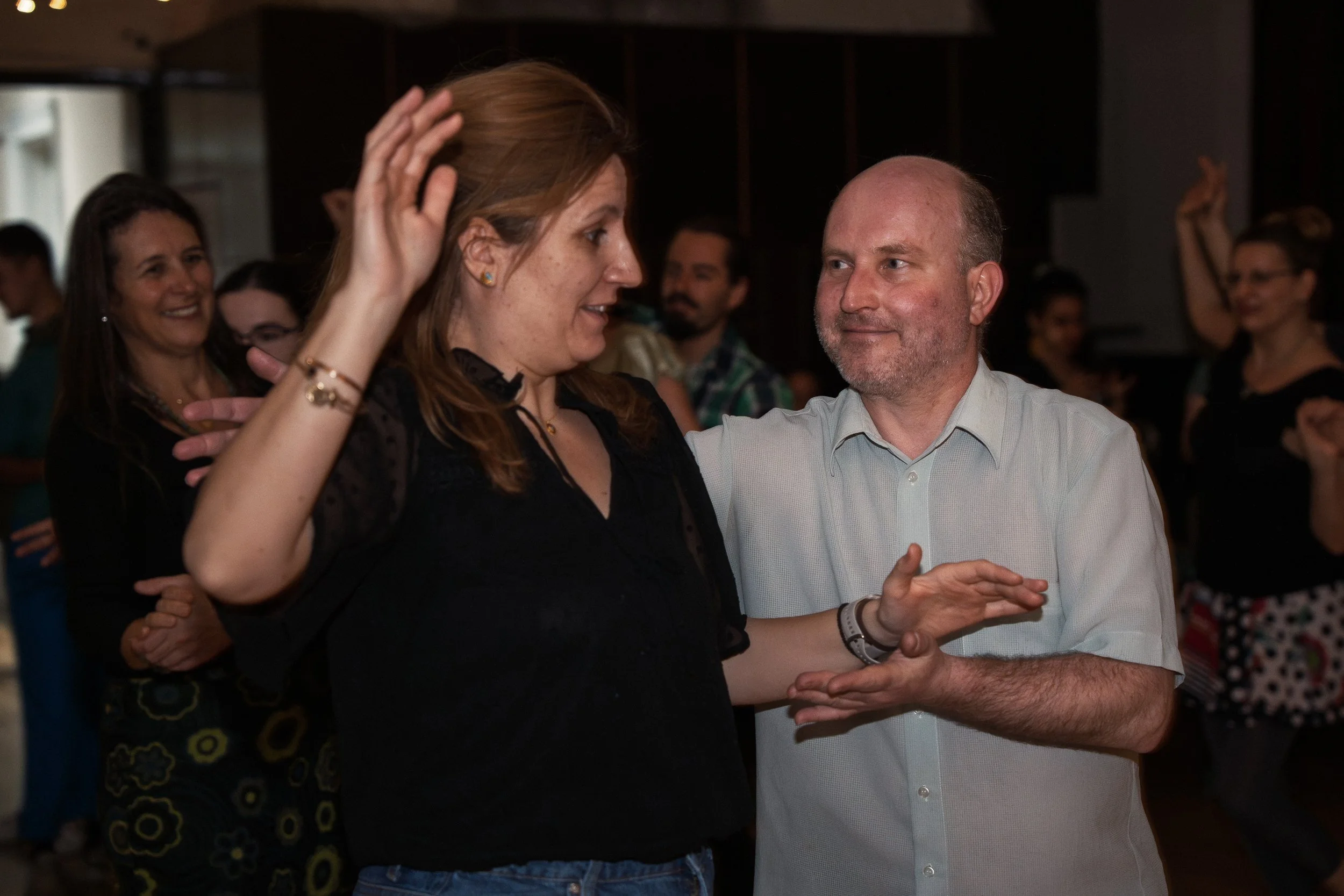 A man and woman dancing together at a social event, with other people smiling and enjoying themselves in the background.