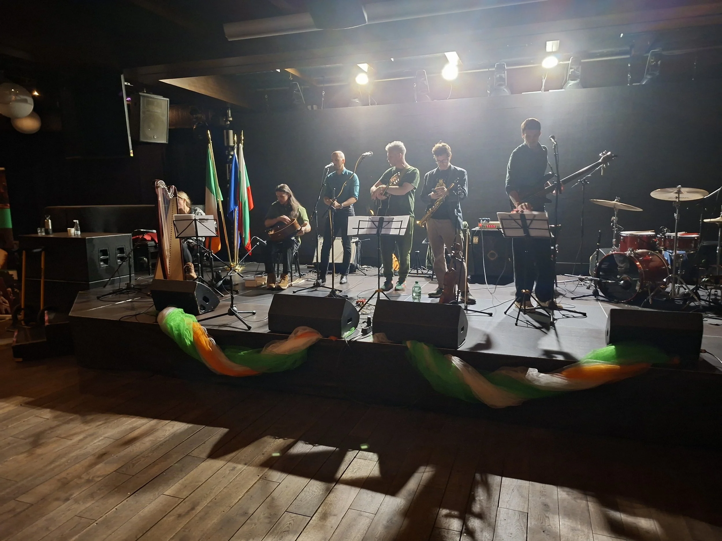 A group of musicians performing on a stage in a dimly lit venue, with bright stage lights shining down. The band includes several members playing instruments such as guitars, saxophone, drums, and a keyboard. There are flags and sound equipment on st