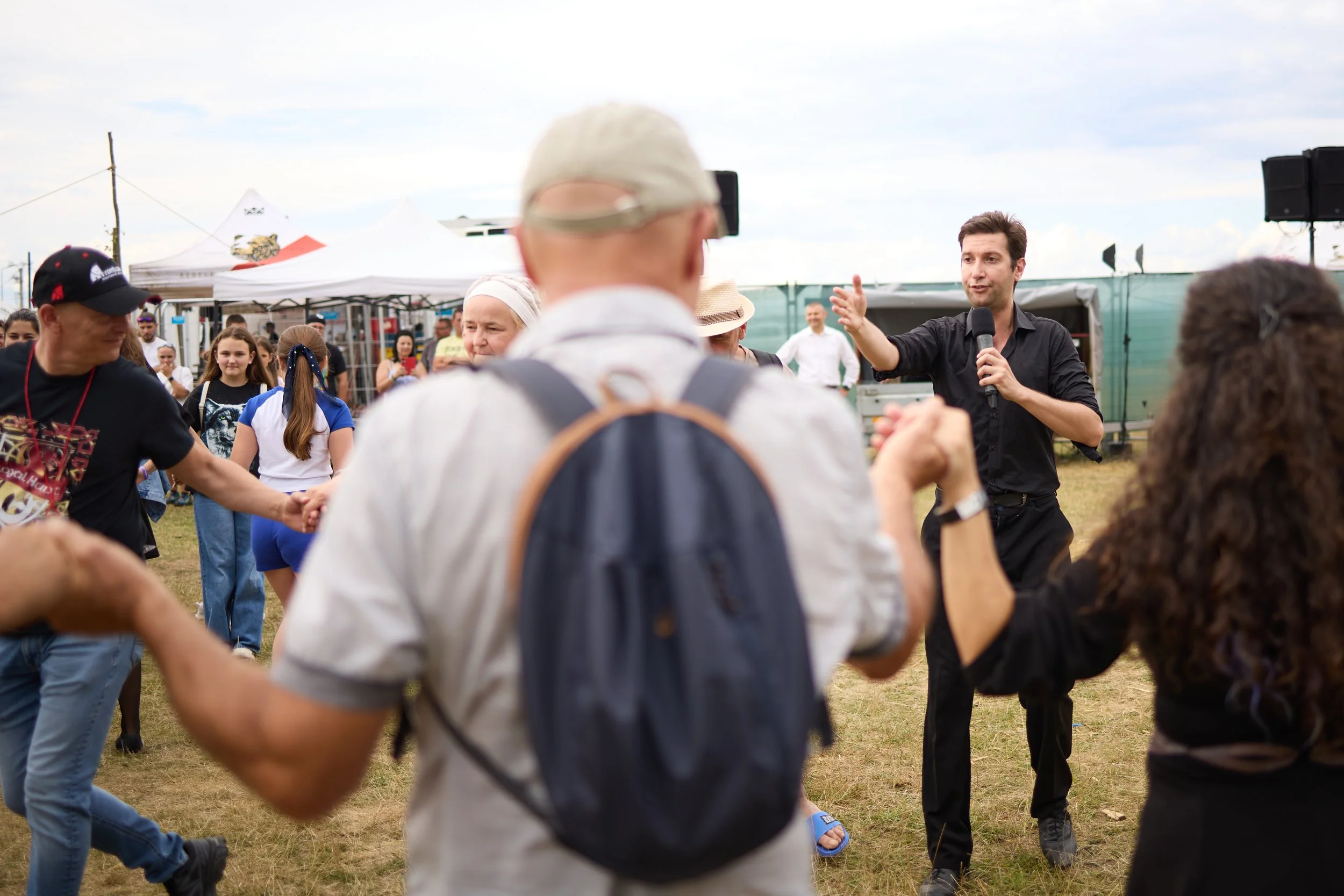 A man in a black shirt speaks to a crowd at an outdoor event. The crowd is diverse, and some people are dancing in a circle. Tents and a stage are visible in the background.