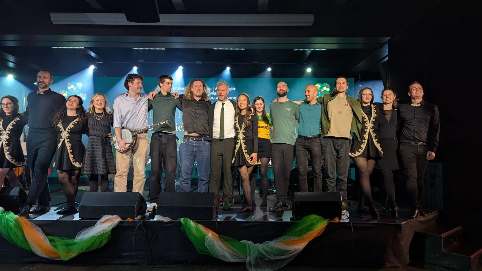 Group of people standing on a stage after a performance or event, some holding musical instruments, with stage lighting and a blue backdrop.
