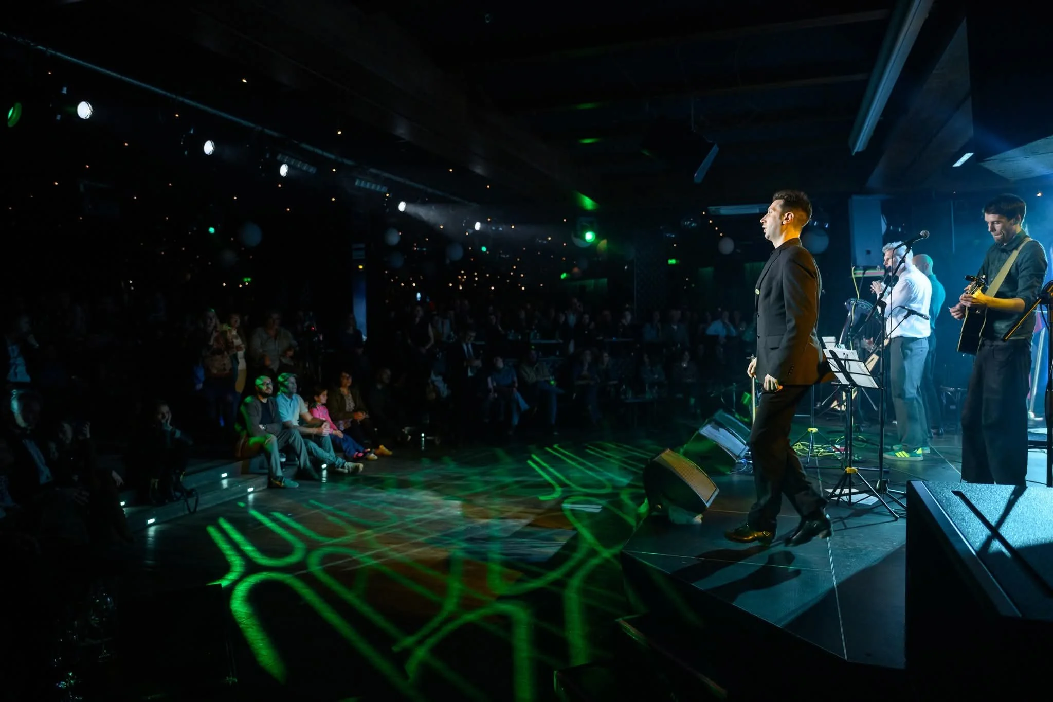 Musicians perform on stage at a concert or event with audience seated in front. Stage lighting includes green lights and spotlights creating a vibrant atmosphere.