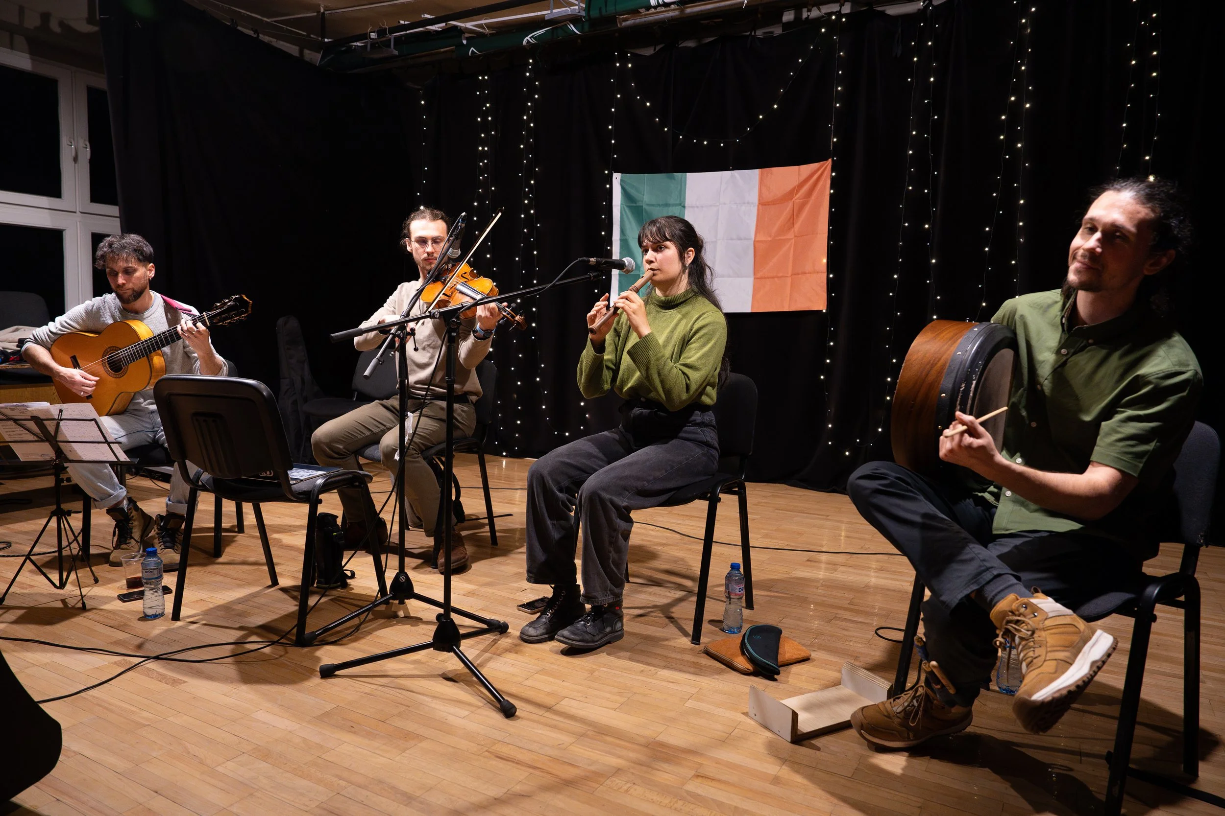 A group of five musicians performing on stage, with two playing guitars, one playing a violin, one playing a wind instrument, and one sitting with a large drum. Behind them is an Irish flag and a backdrop of string lights.