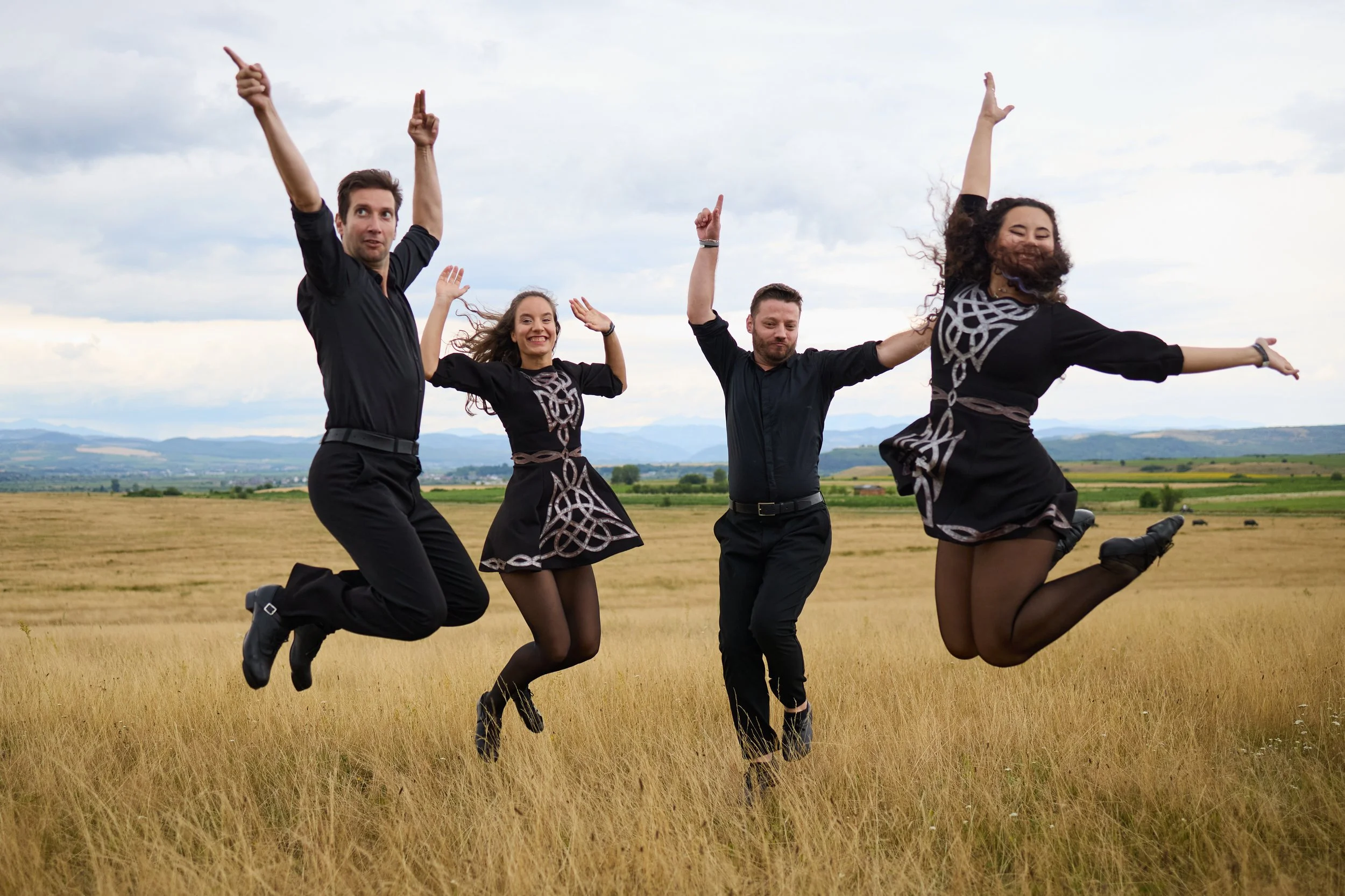 Four people jumping in a grassy field with mountains in the background during cloudy weather, all dressed in black and smiling.