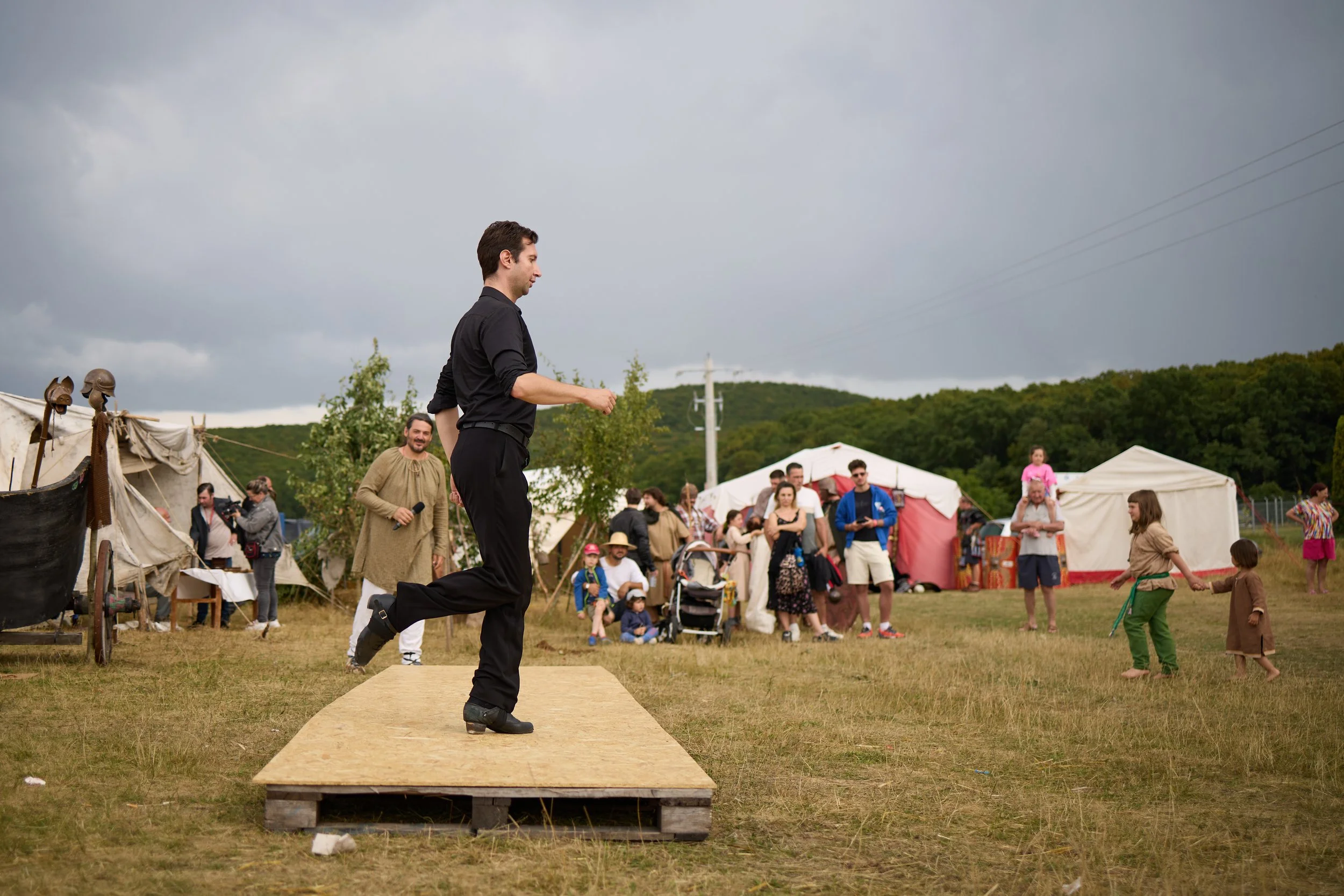 A man dressed as a Roman or medieval soldier is kneeling on a platform, preparing to stand or perform, at an outdoor event with tents and a crowd of people including children and adults, under a cloudy sky.