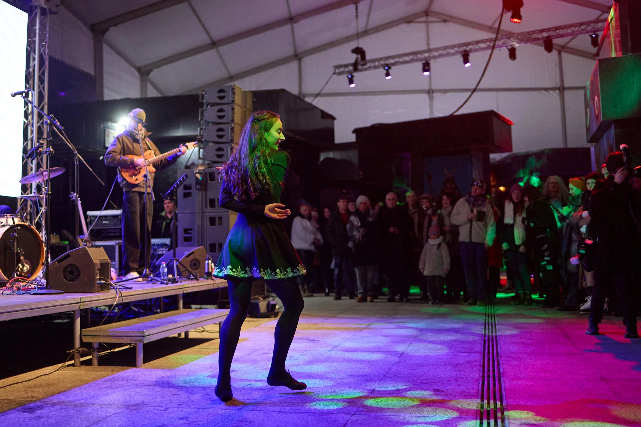 A woman dancing in a darkened event tent with stage lights, in front of a live musician playing guitar. Audience members observe from the background.