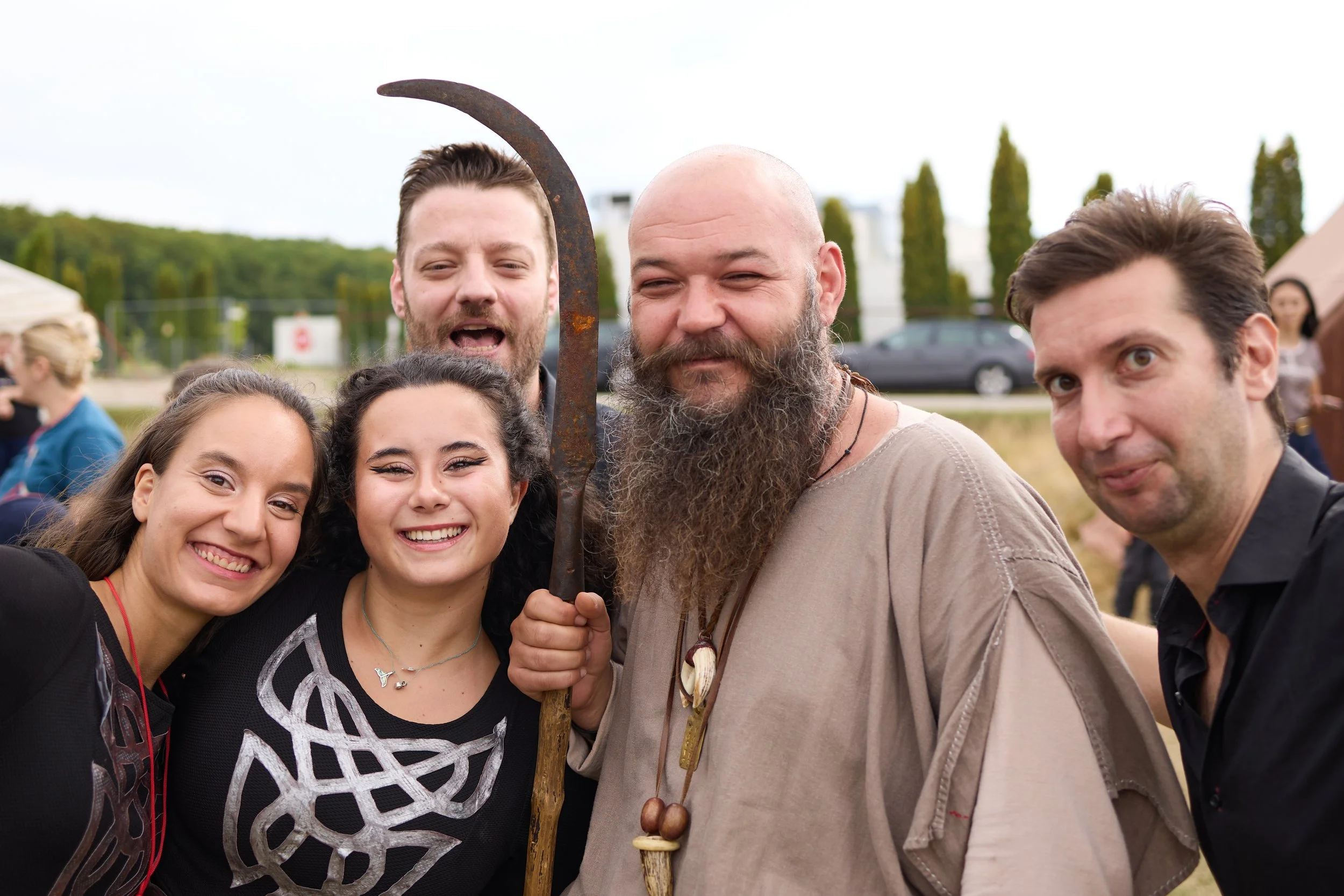Group of people at an outdoor event, smiling, with a man holding a rusted sickle and wearing a necklace made of shells, with trees and tents in the background.