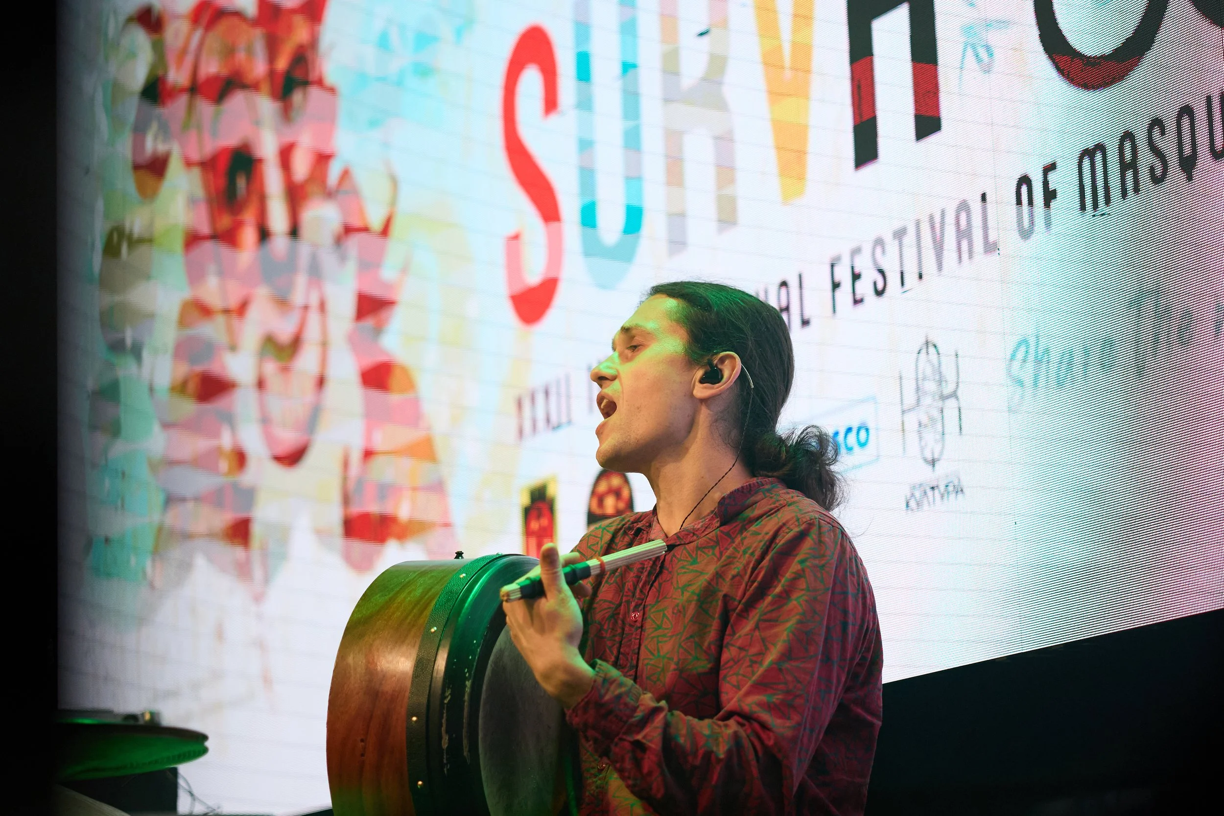 A person with long dark hair playing a hand drum at a festival or event, with a colorful screen in the background displaying text and logos.