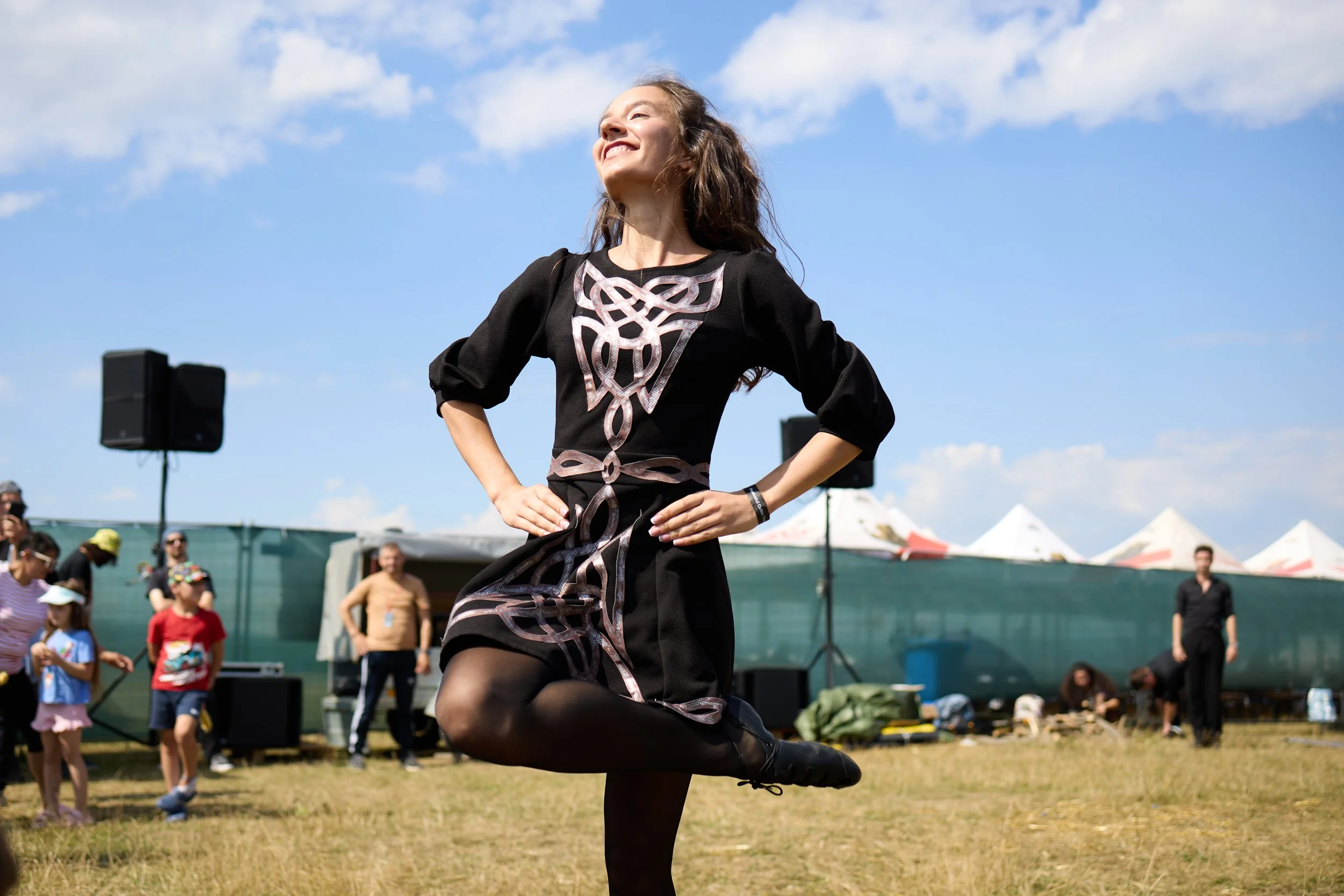 A woman in a black dress with Celtic knot designs is performing a dance outdoors, standing on one leg with a joyful expression, while a crowd watches on a bright, sunny day with blue skies and white clouds.