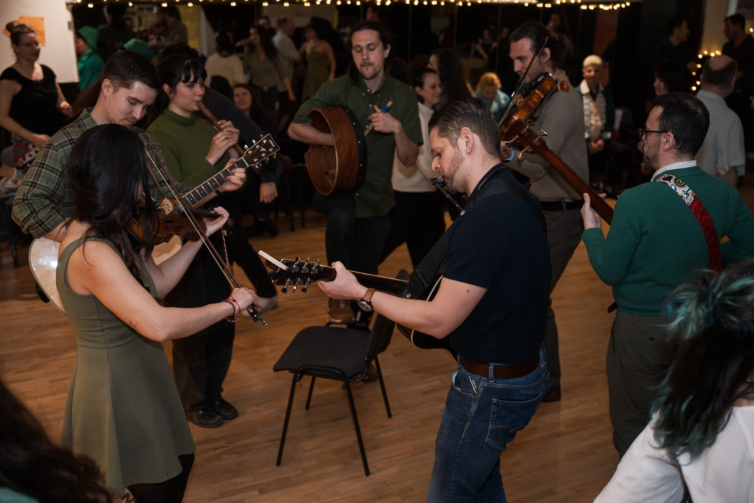 People playing musical instruments together at an indoor gathering, with an audience in the background.