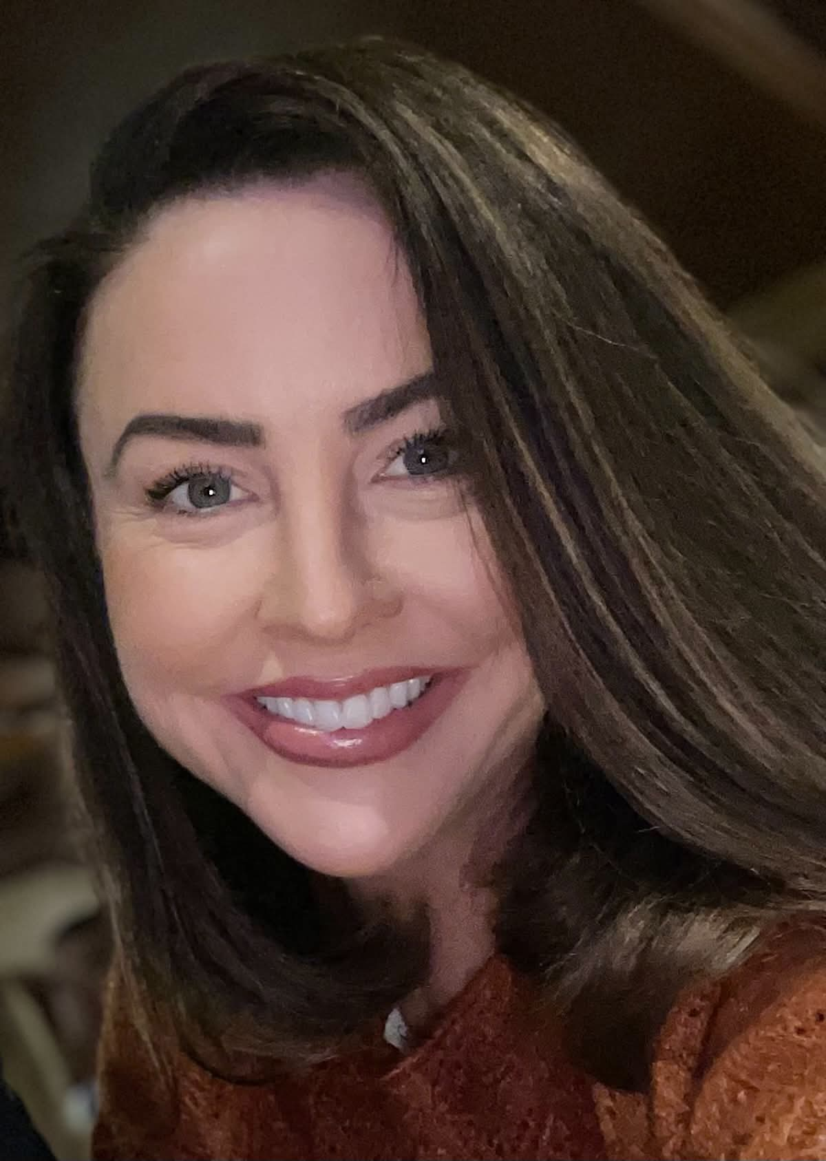 A smiling woman with long brown hair, blue eyes, and wearing a rust-colored top, looking directly at the camera.