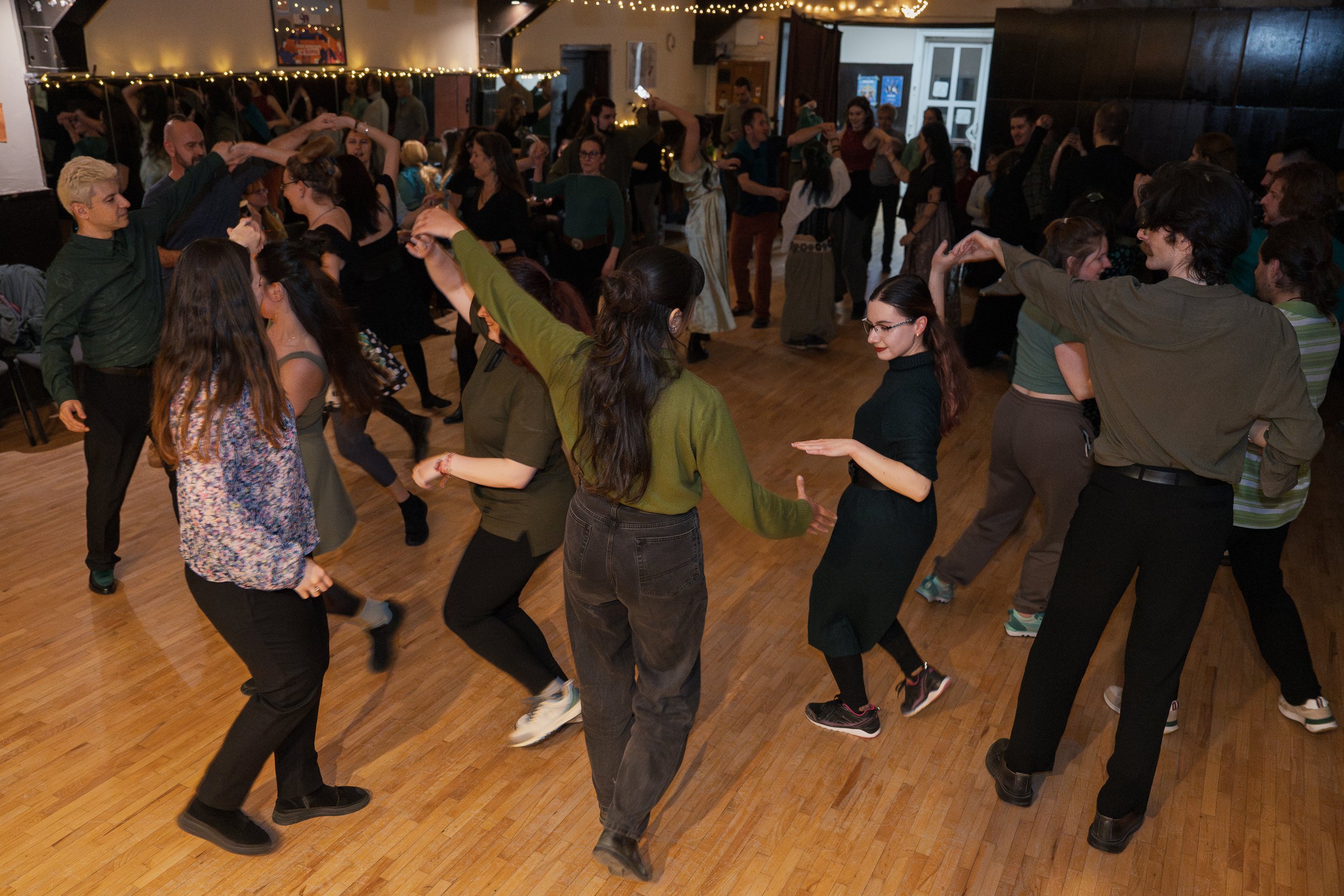 People dancing in a crowded indoor space with wooden floors, string lights, and a mirror wall.