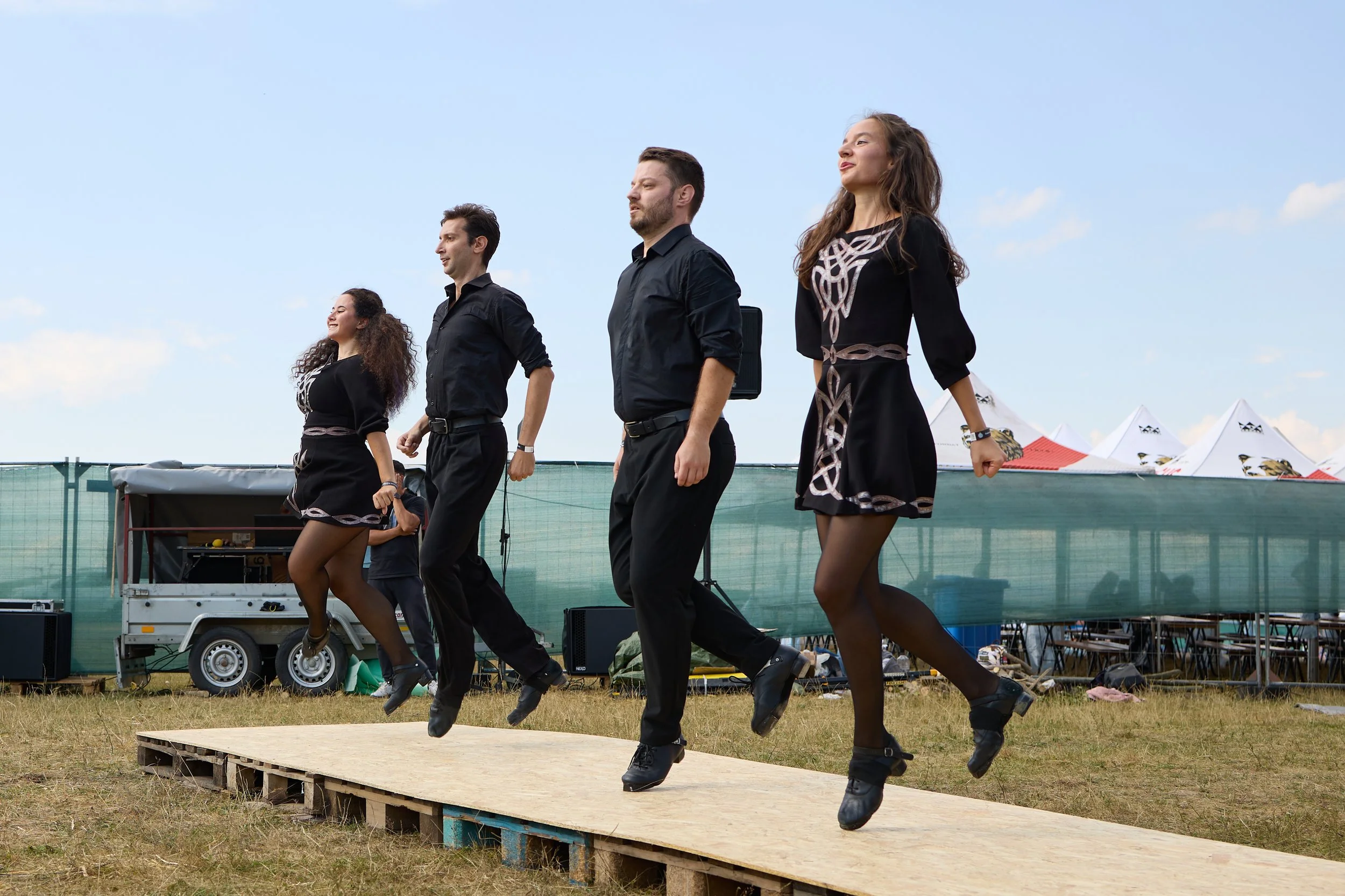 Group of five dancers performing a coordinated dance routine outdoors on a wooden stage, dressed in black outfits with intricate patterns, under a clear blue sky with tents in the background.