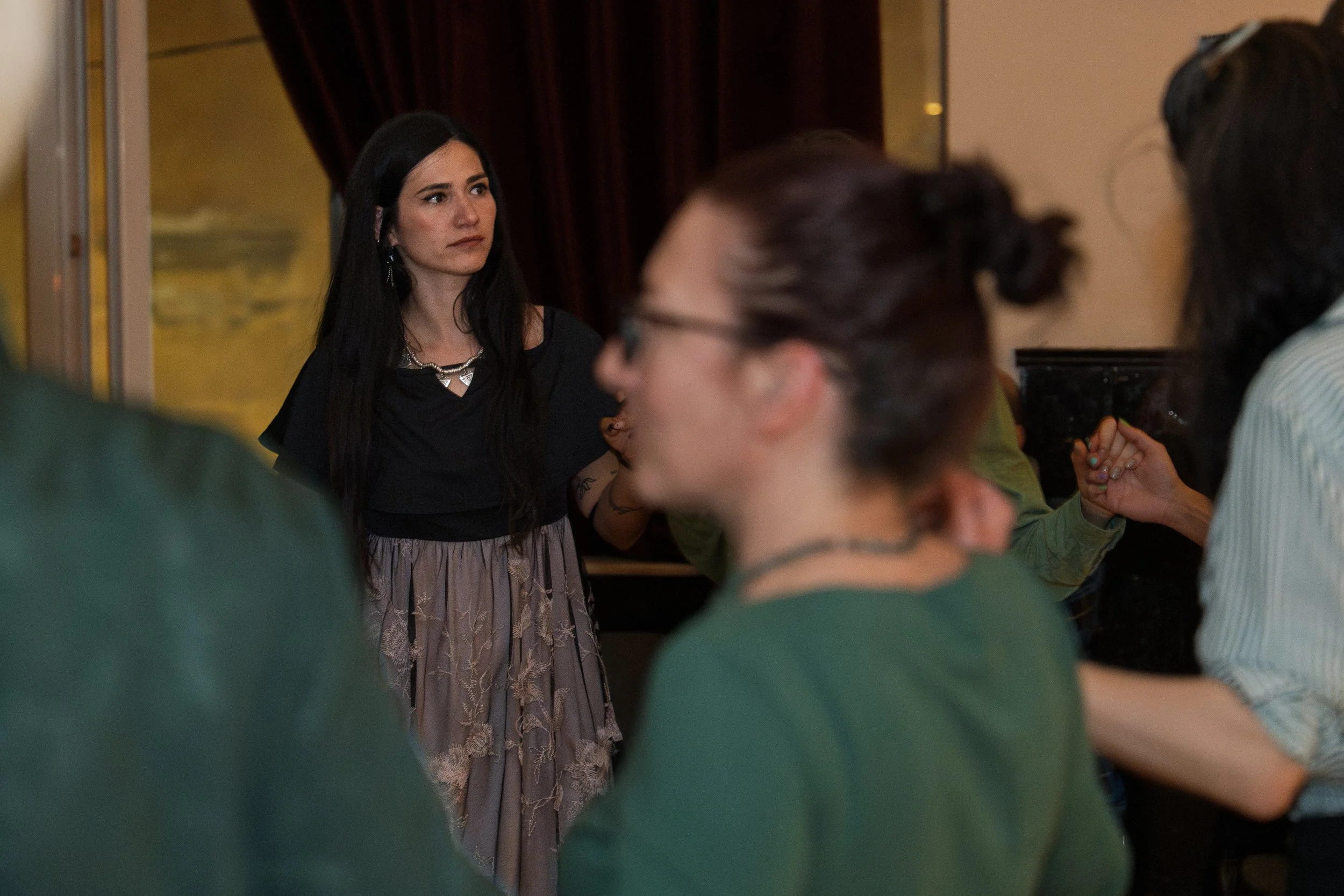 A woman with long black hair, wearing a black top and a patterned gray skirt, standing and looking to her left at an event with several people surrounding her.