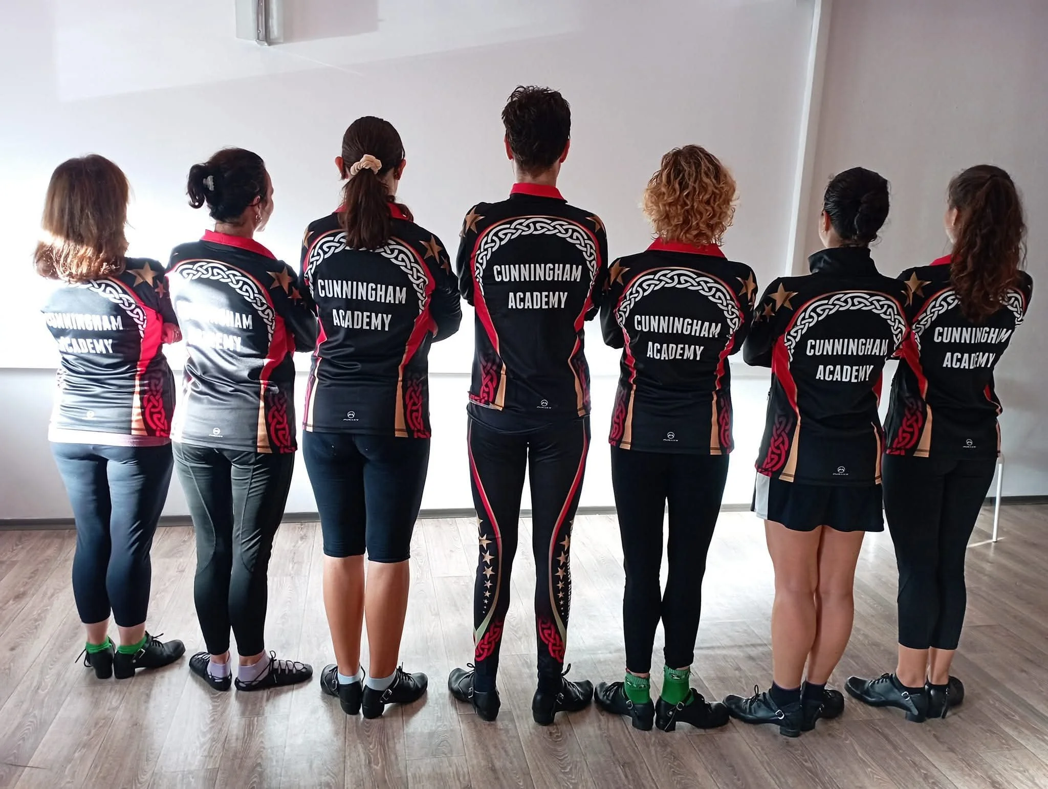 Seven people standing in a row with their backs to the camera, wearing matching black and red sports jerseys with 'CUNNINGHAM ACADEMY' printed on the back, in a room with wooden flooring and plain white walls.
