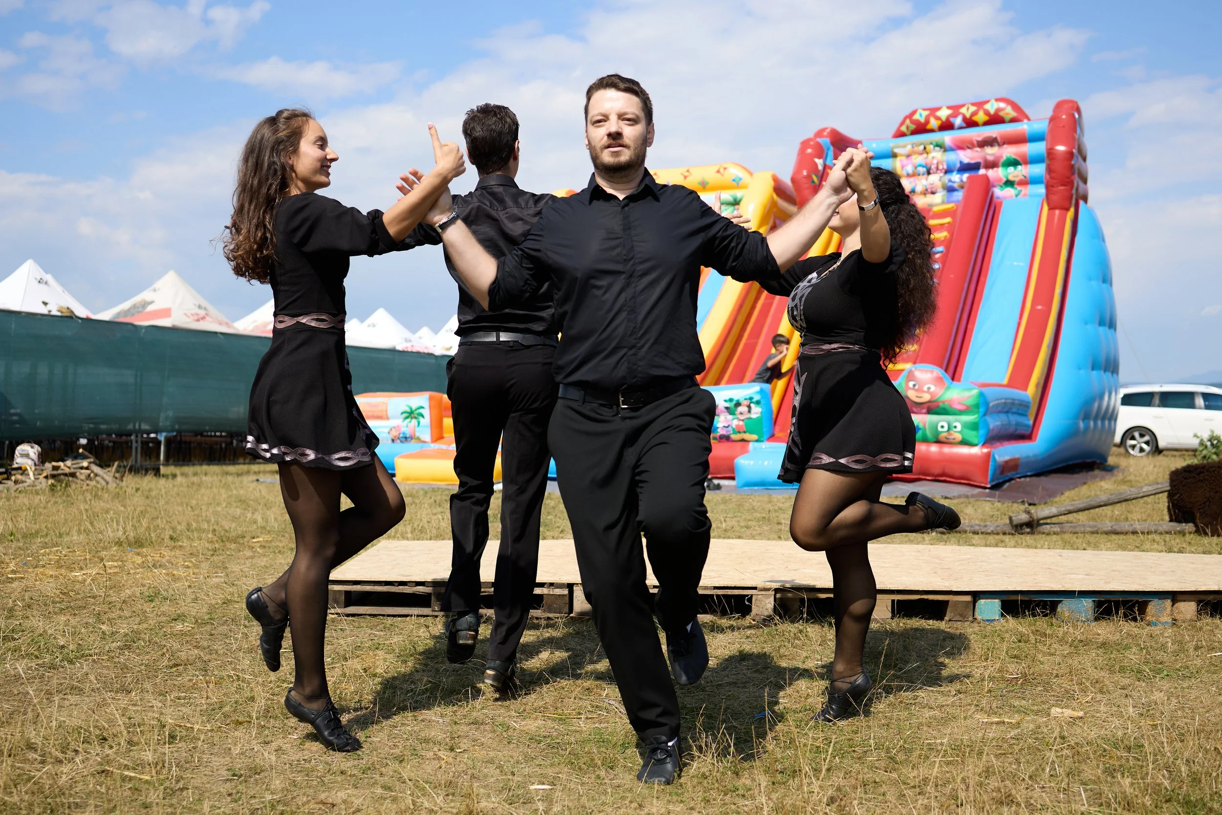 Four people, dressed in black, dancing outdoors in front of an inflatable slide with cartoon characters, during daytime under a blue sky with some clouds.
