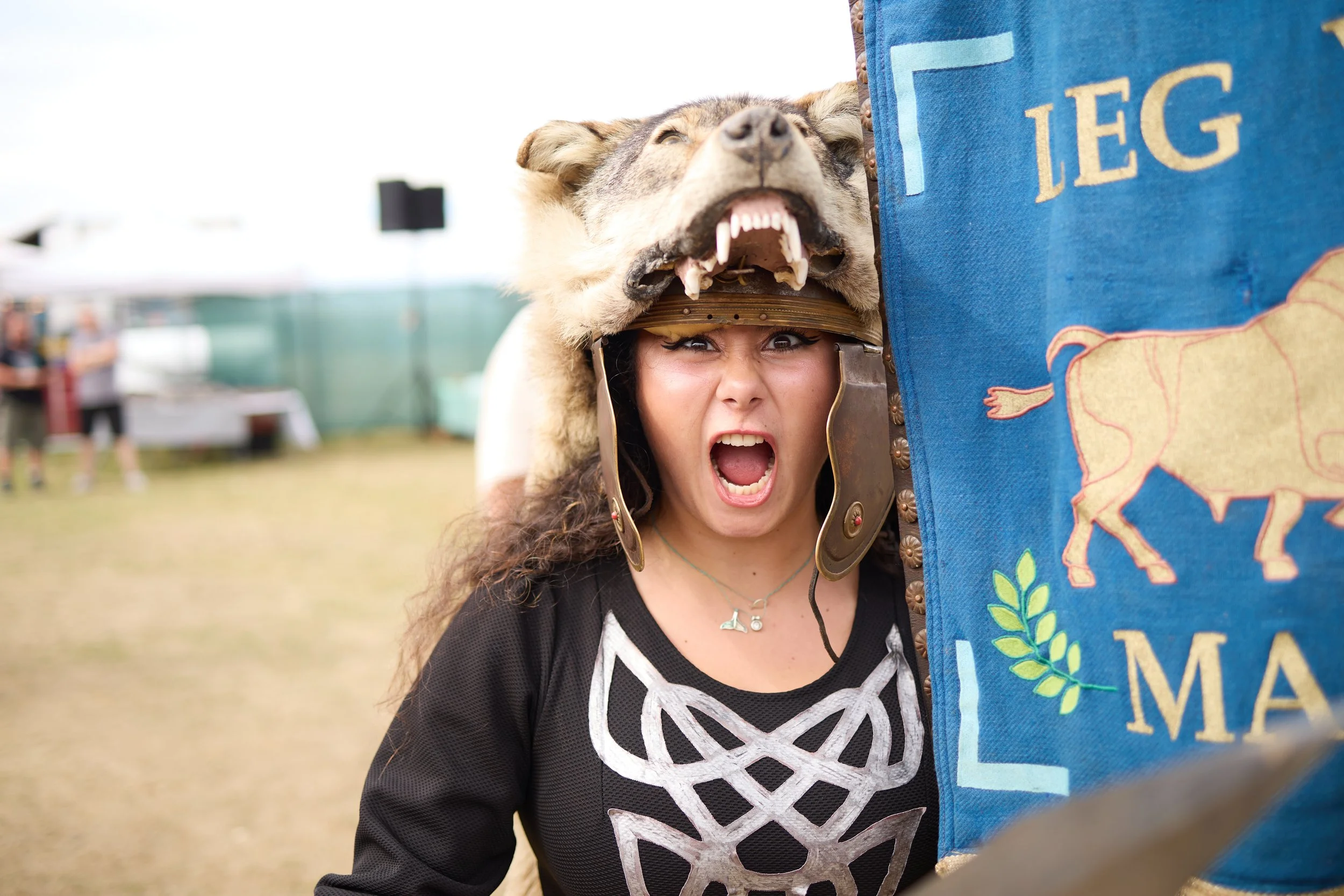 A woman wearing a wolf headdress and a black shirt with white lace is standing outdoors, partially behind a blue banner with a yellow bison and green leaves. She is shouting or cheering with her mouth open and her eyes wide.