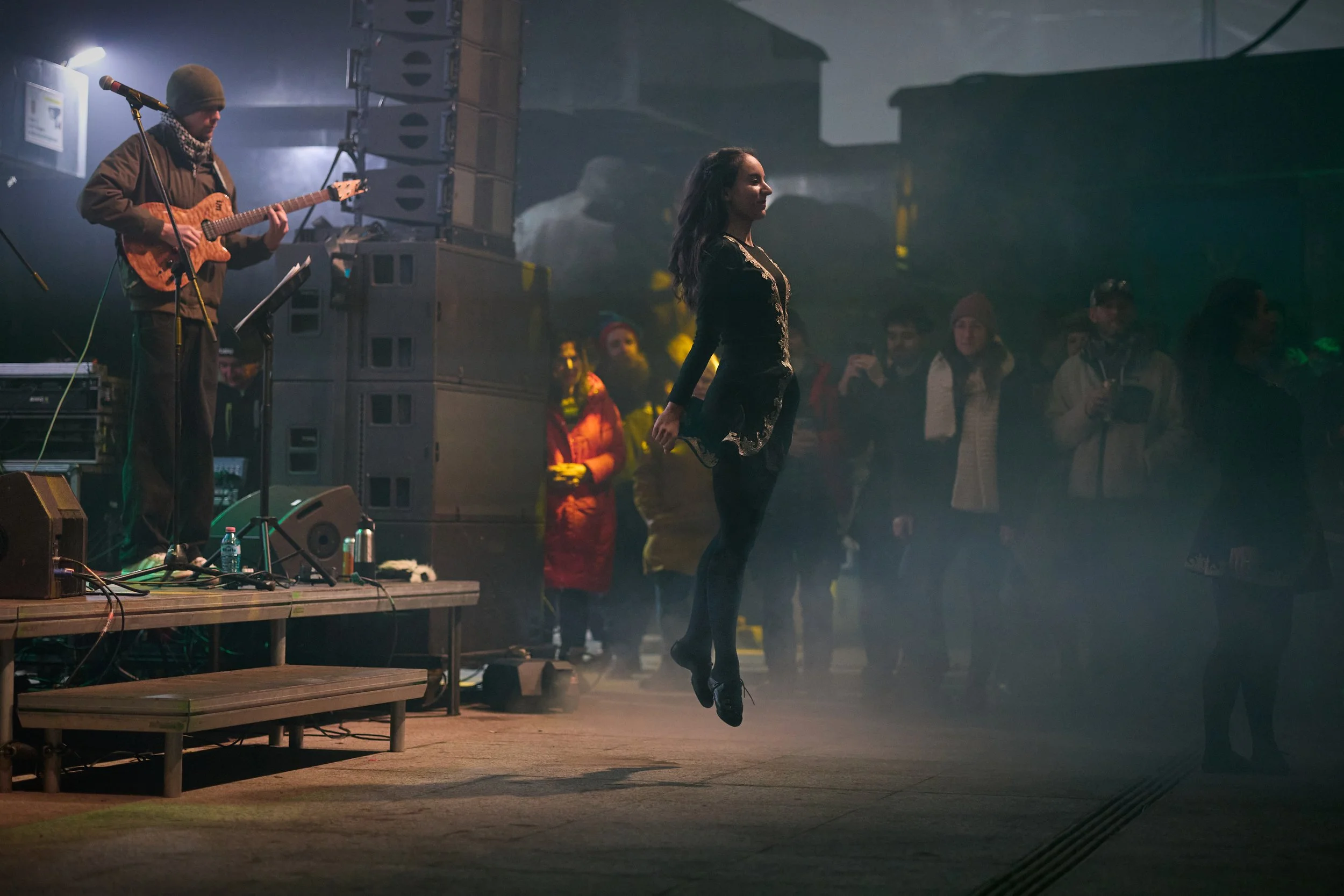 A woman in black pants and a dark top with gold embroidery jumps in the air during a performance, with a man in a beanie and scarf playing an electric guitar on stage. The audience watches in a dimly lit venue with speakers and equipment visible.