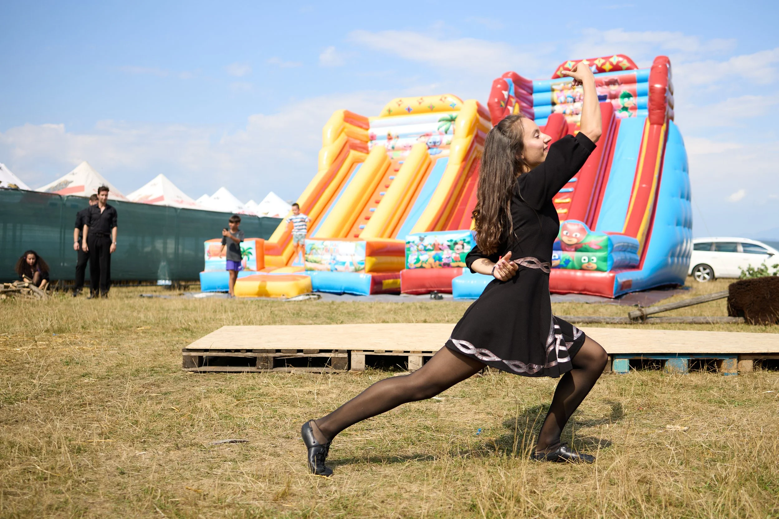 A woman in a black dress and black heels performing a dance move in front of an inflatable slide at an outdoor event. The inflatable slide has bright colors and cartoon characters. There are children and adults in the background, some standing near t
