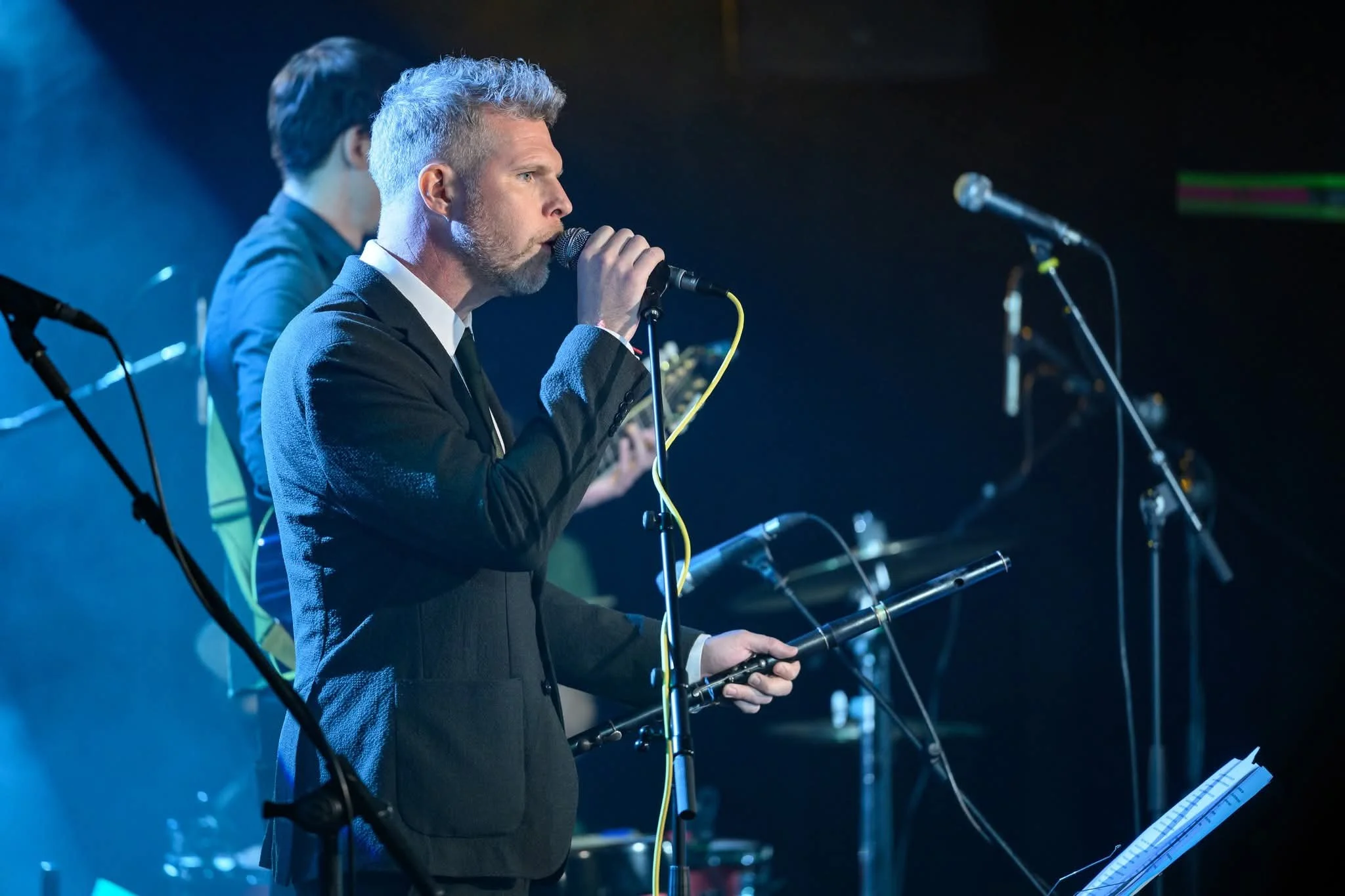 A man in a suit singing into a microphone during a performance on stage, with a keyboardist or musician visible in the background.