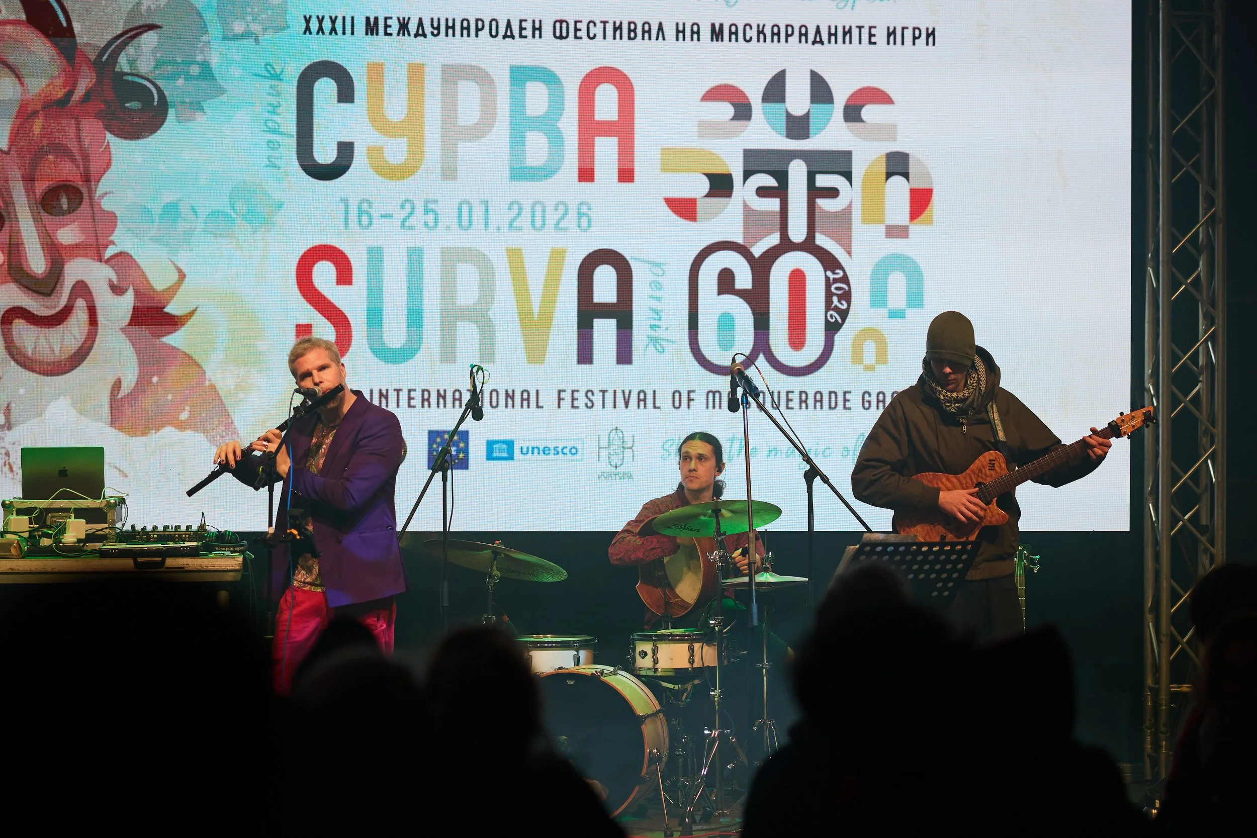 A band performing live on stage at an international festival of masquerade games, with a large colorful banner in the background displaying event details and logos.