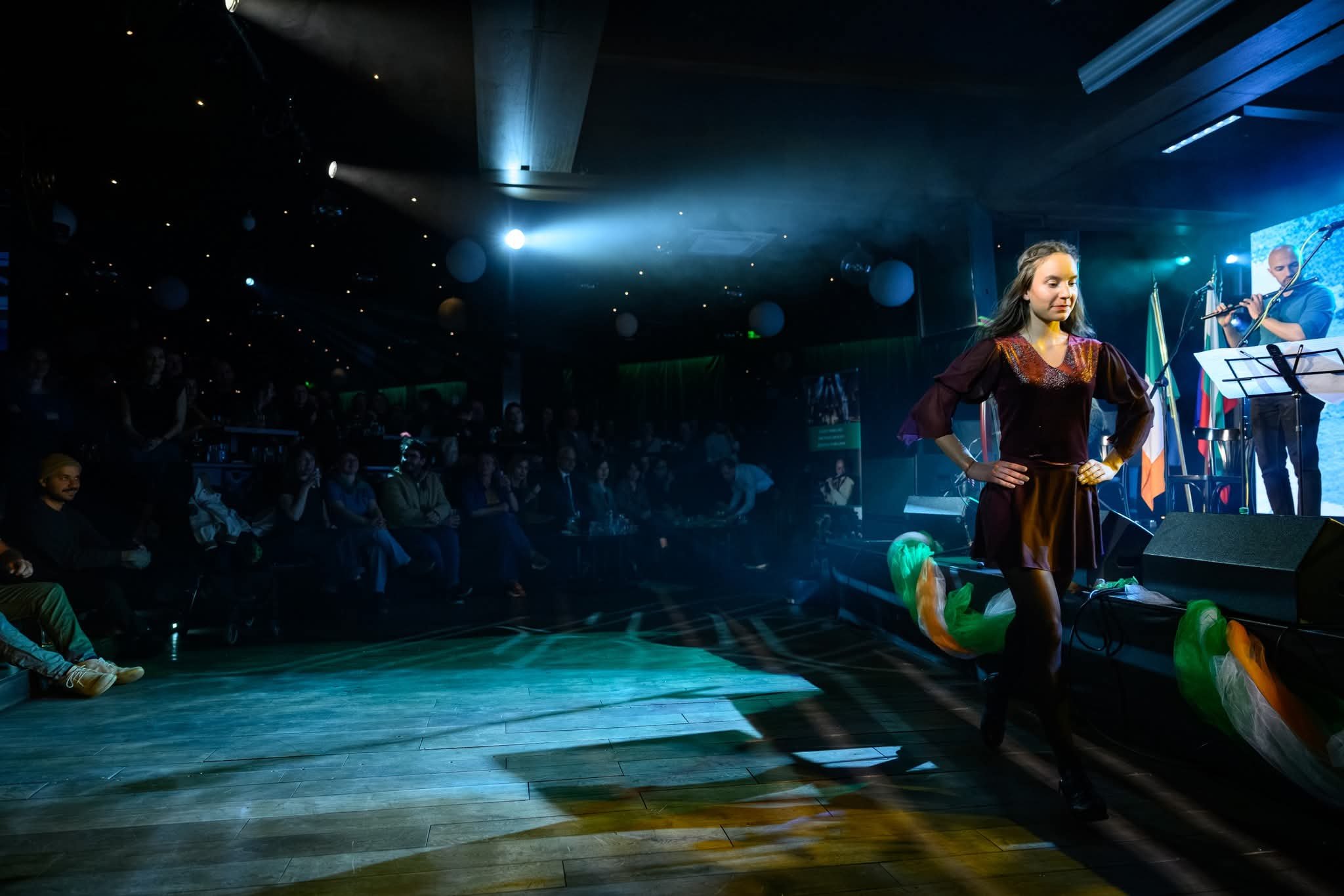 A woman posing confidently on a stage at a live performance, with a musician playing guitar behind her and audience seated in the dark watching. The stage has colorful lighting and decorations.