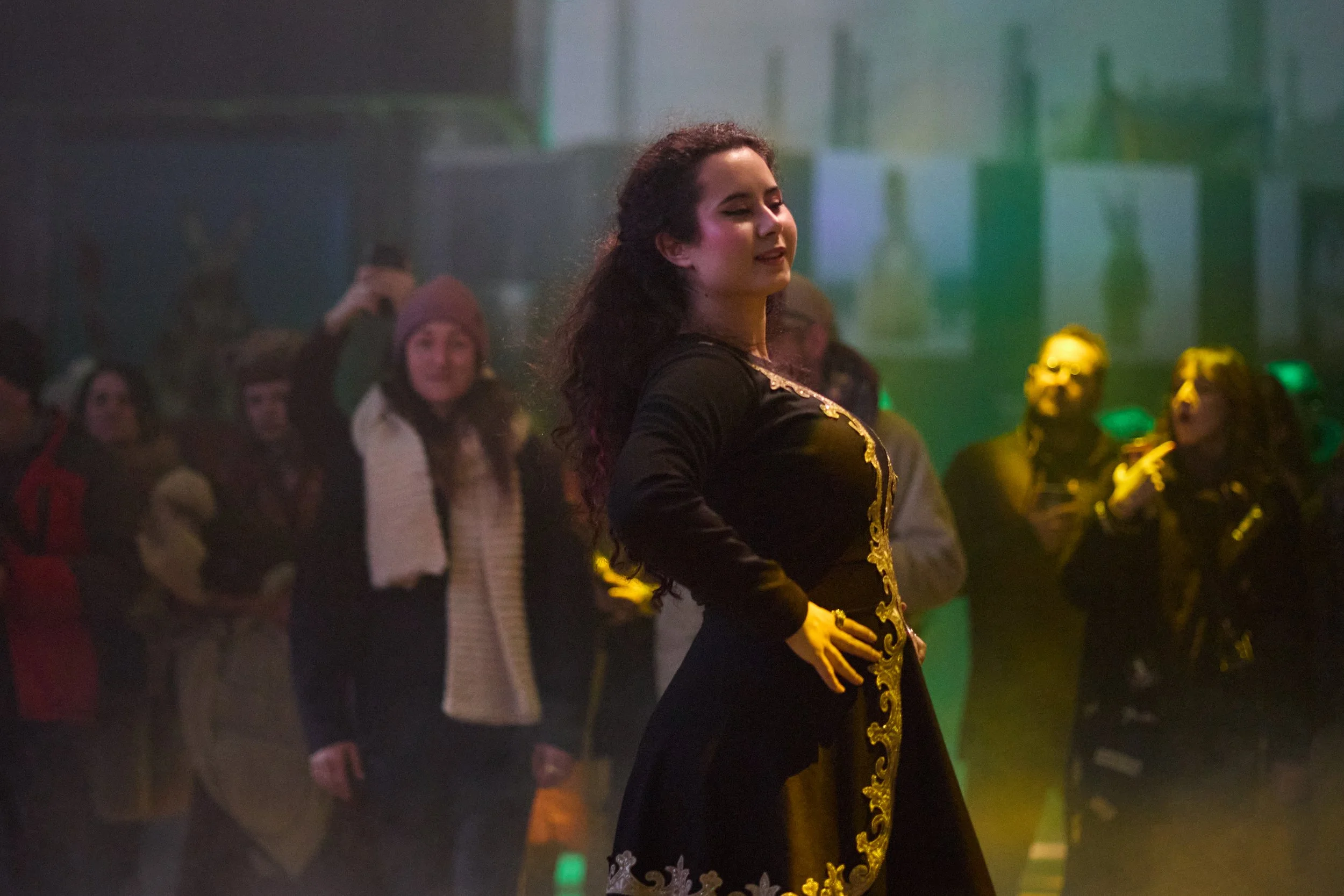 A woman in a black dress with gold embroidery dancing at a social gathering with people watching and taking pictures in a dimly lit room.